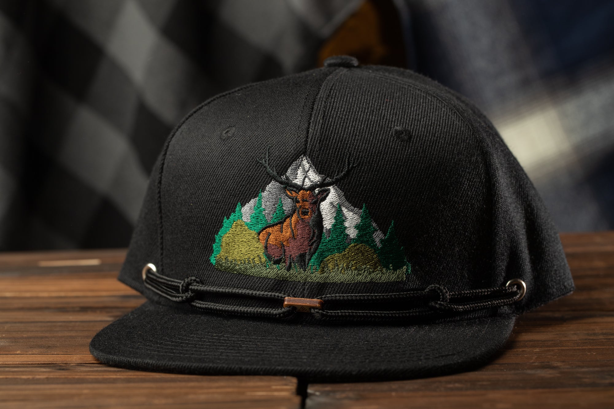 A black baseball cap with an embroidered design of a deer standing in a forest on a wooden surface.