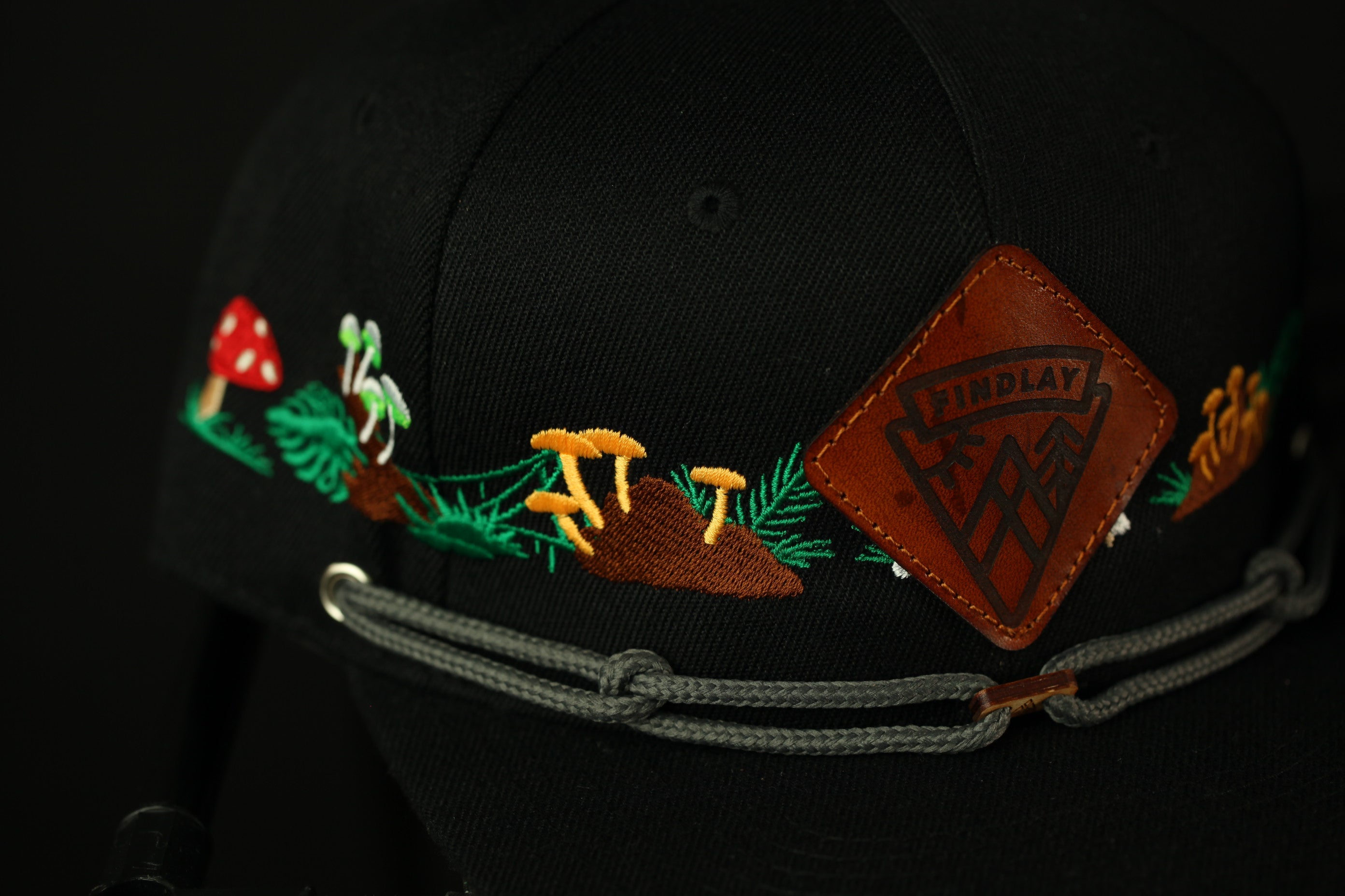 a close-up view of a black baseball cap with a decorative embroidered design featuring mushrooms, leaves, and other natural elements.