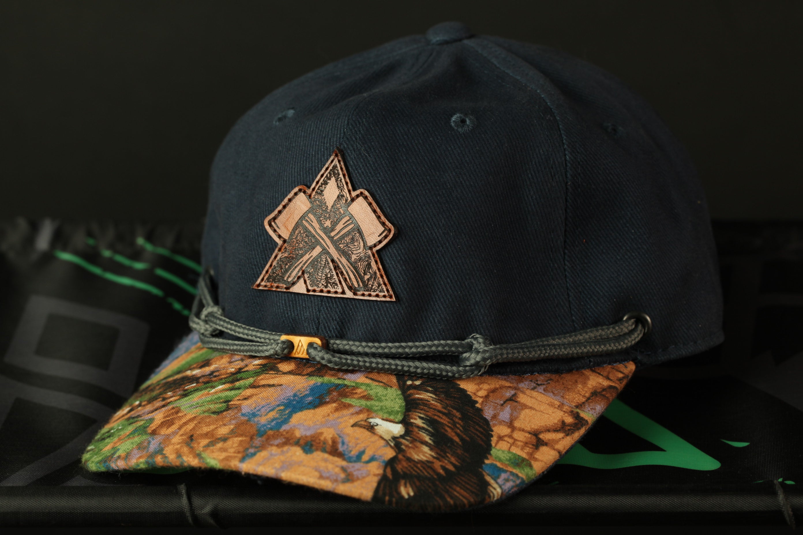 A dark blue baseball cap with a camouflage pattern and a patch on the front, placed on a surface with a green and black patterned background.