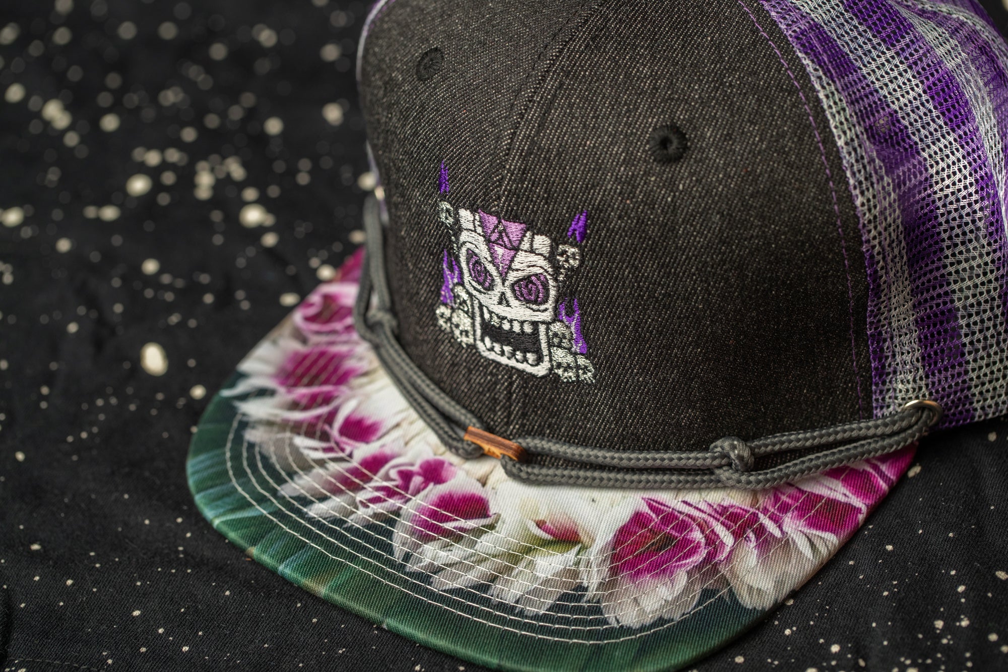 A close-up image of a black baseball cap with a colorful floral pattern and a skull-like design on the front.