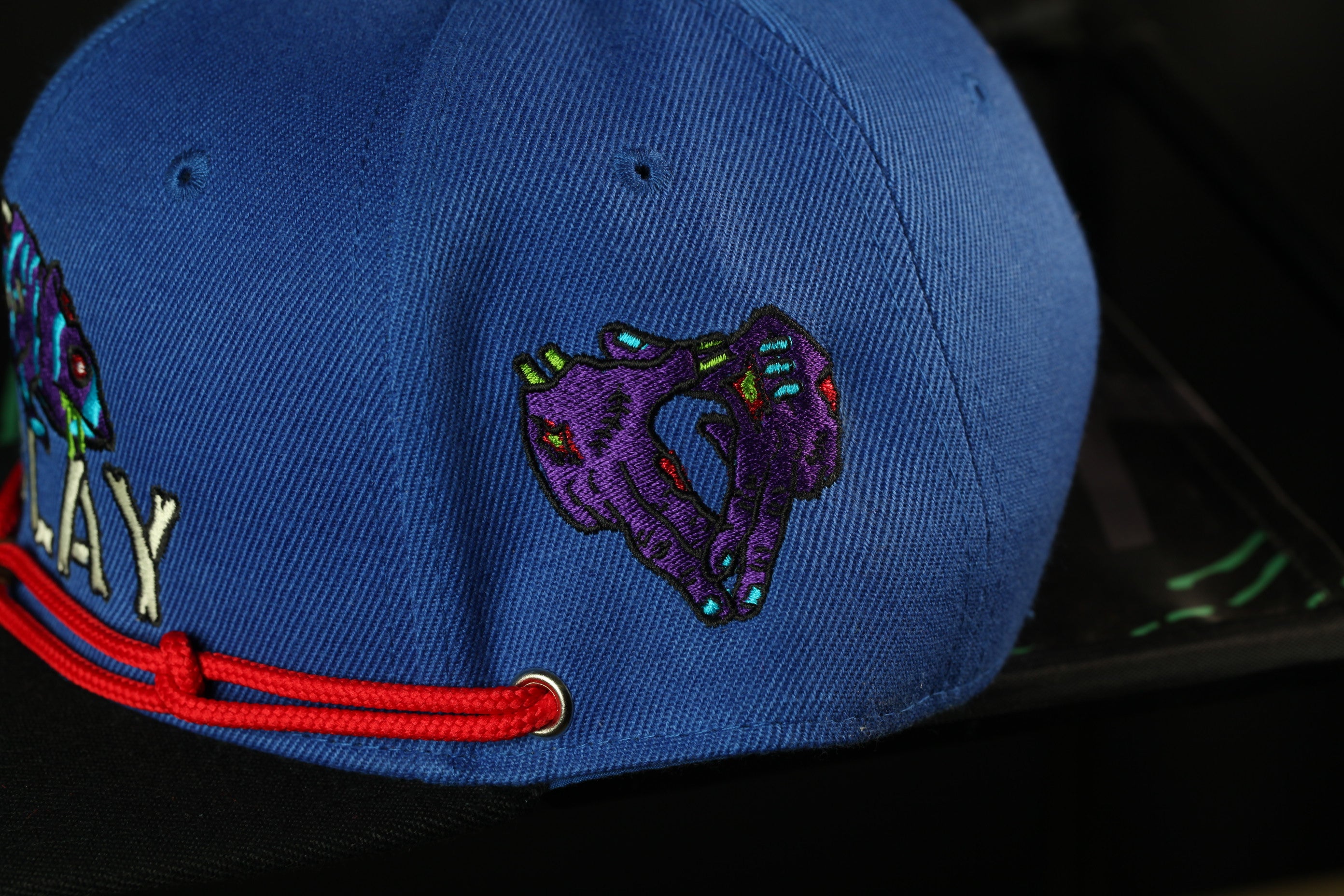 A close-up view of a blue baseball cap with a red string and a colorful, embroidered patch on the front.