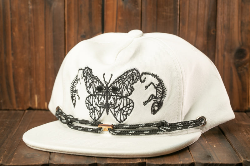 A white baseball cap with a black butterfly design on the front, sitting on a wooden surface.