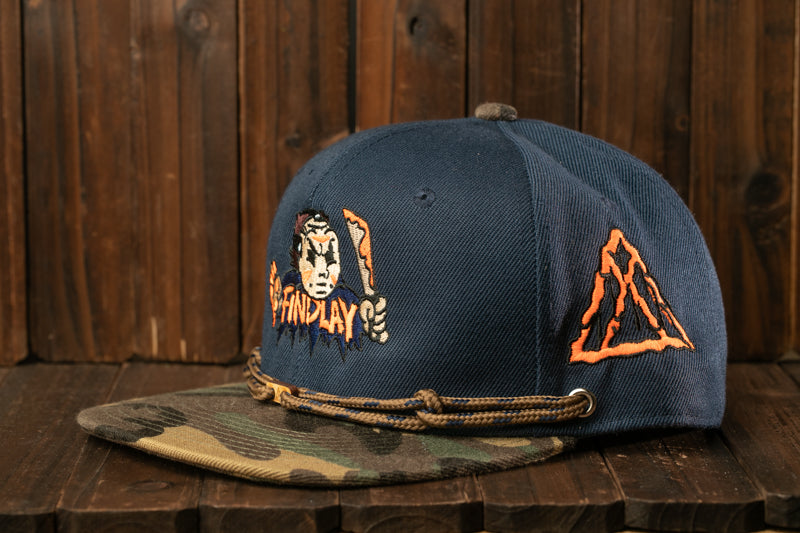 A navy blue baseball cap with a camouflage pattern and a graphic design on the front, placed on a wooden surface.