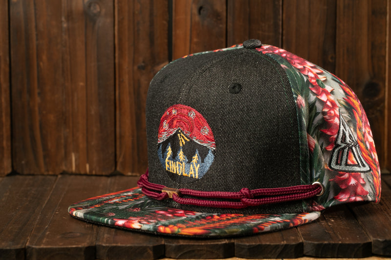 A black baseball cap with a floral pattern and a red band around the brim, placed on a wooden surface.