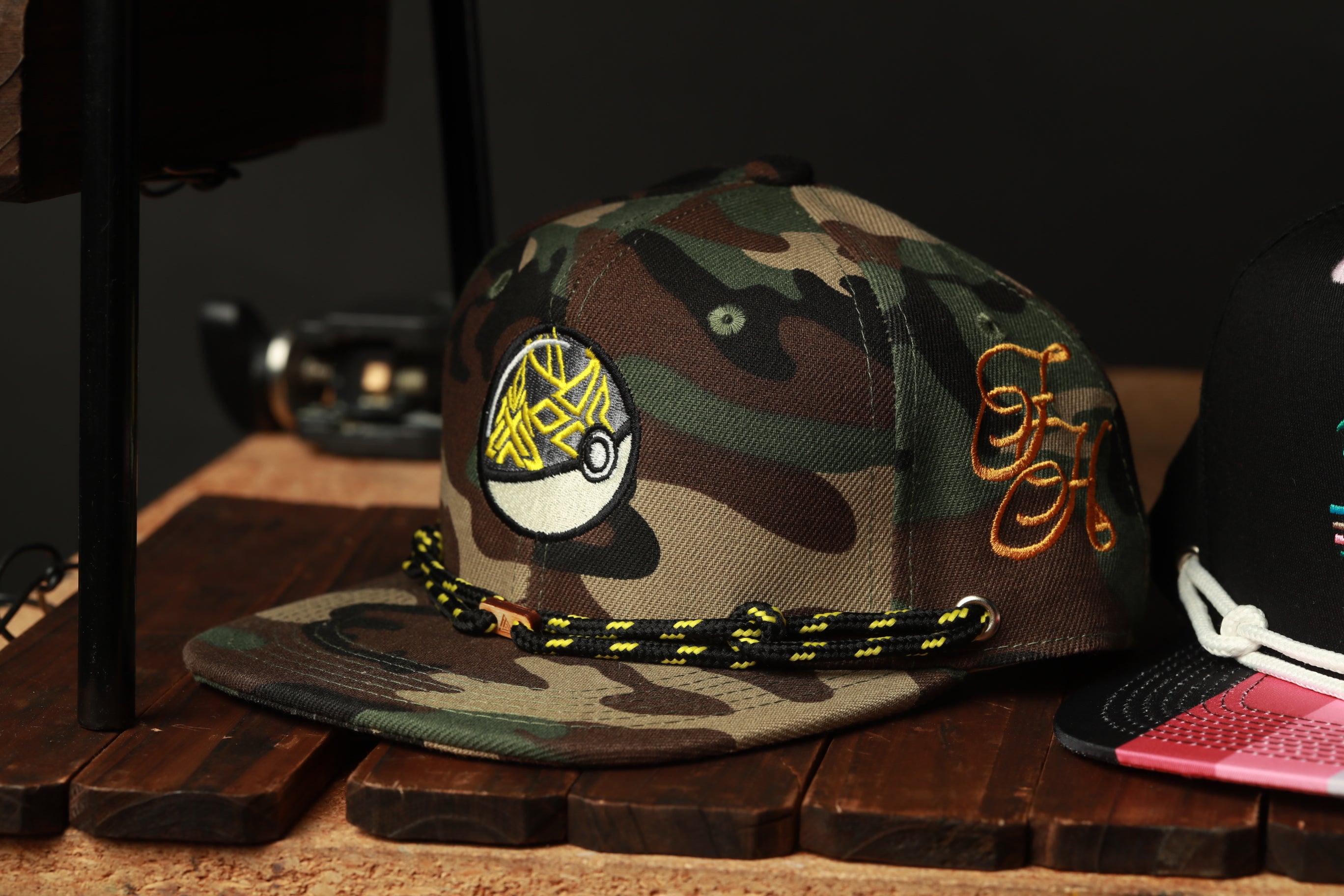 A camouflage baseball cap with a fish design and a black and yellow braided strap is displayed on a wooden surface.