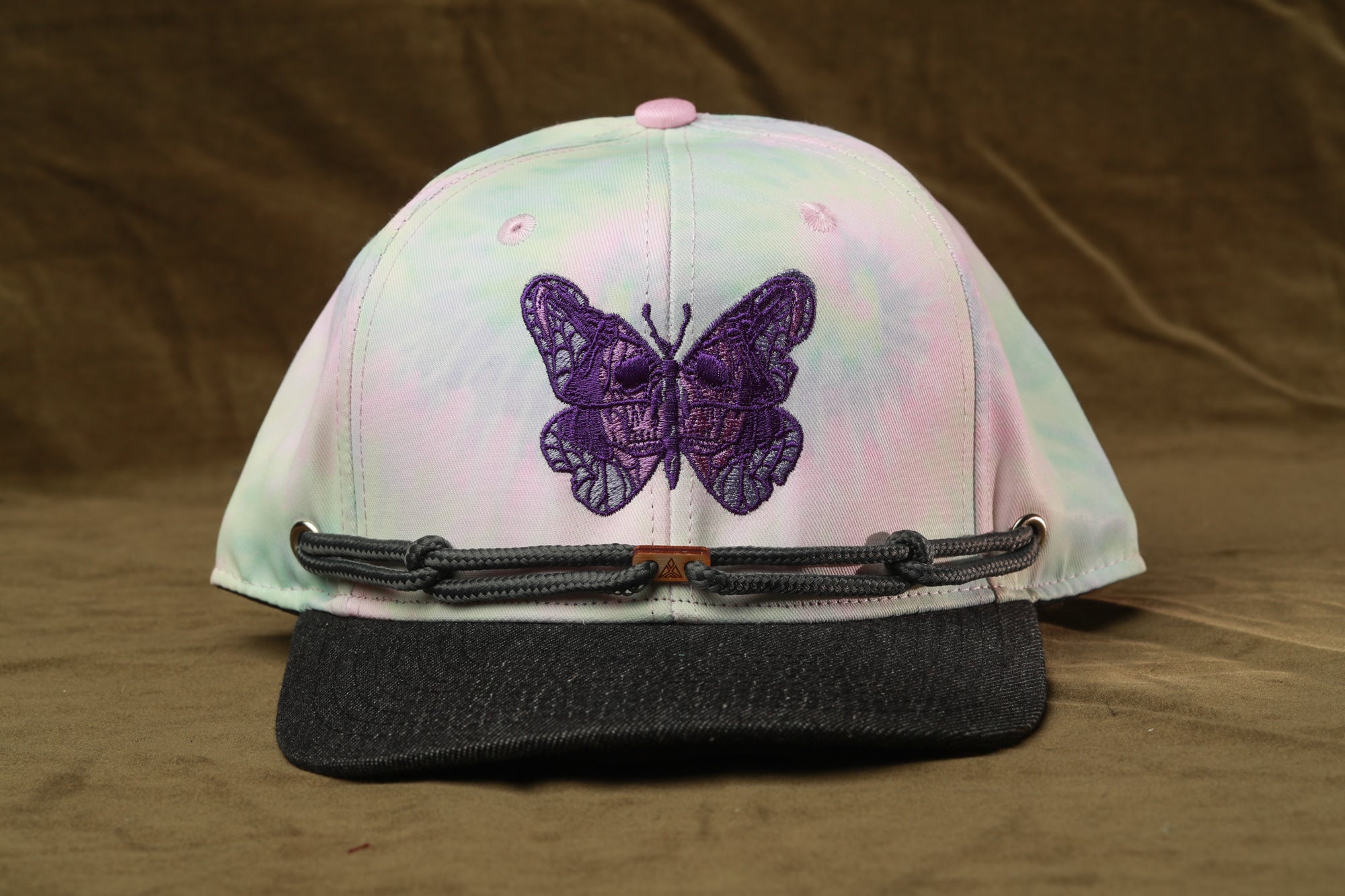 A tie-dye baseball cap with a purple butterfly embroidered on it, resting on a brown surface.