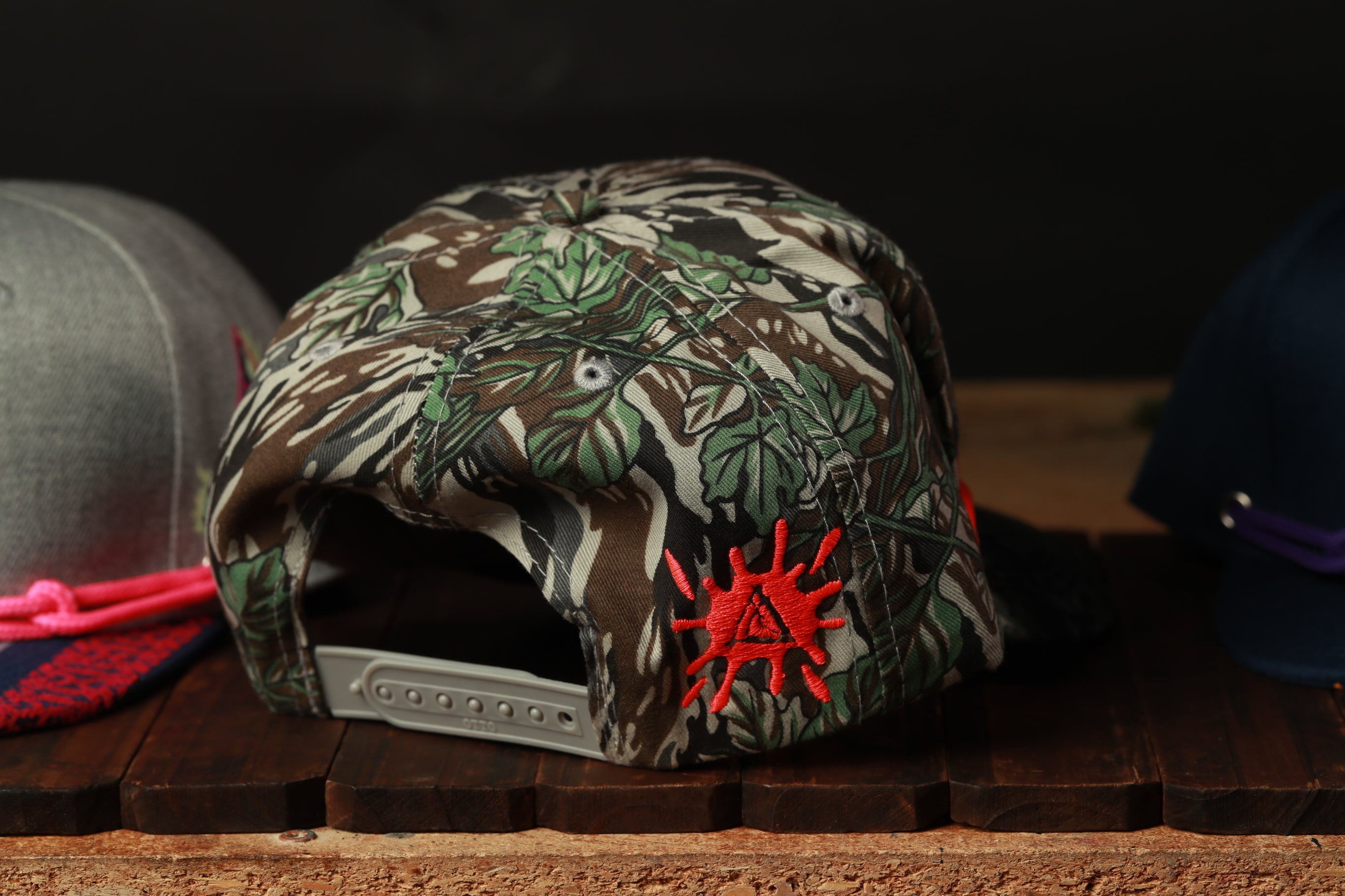 A camouflage baseball cap with a red starburst design is displayed on a wooden surface, with other hats visible in the background.