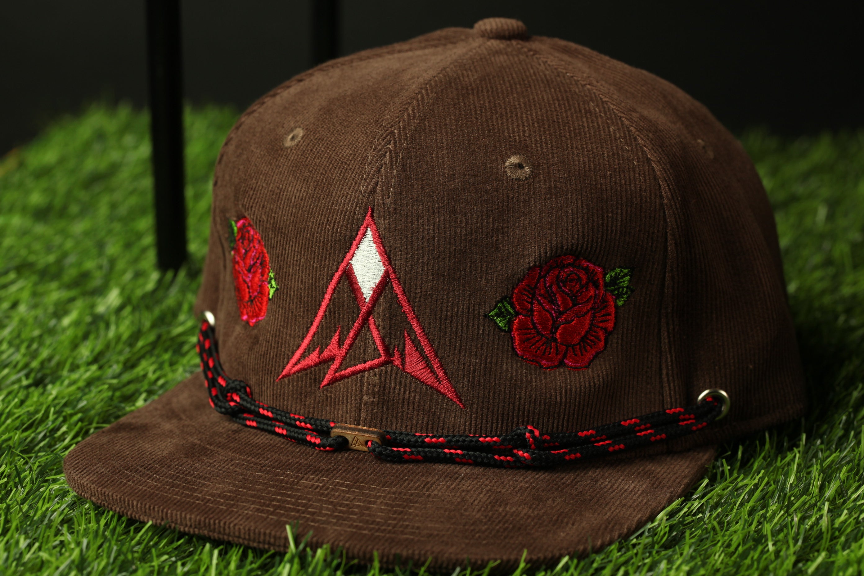 A brown baseball cap with red rose embroidery and a triangular logo on the front, sitting on a grassy surface.