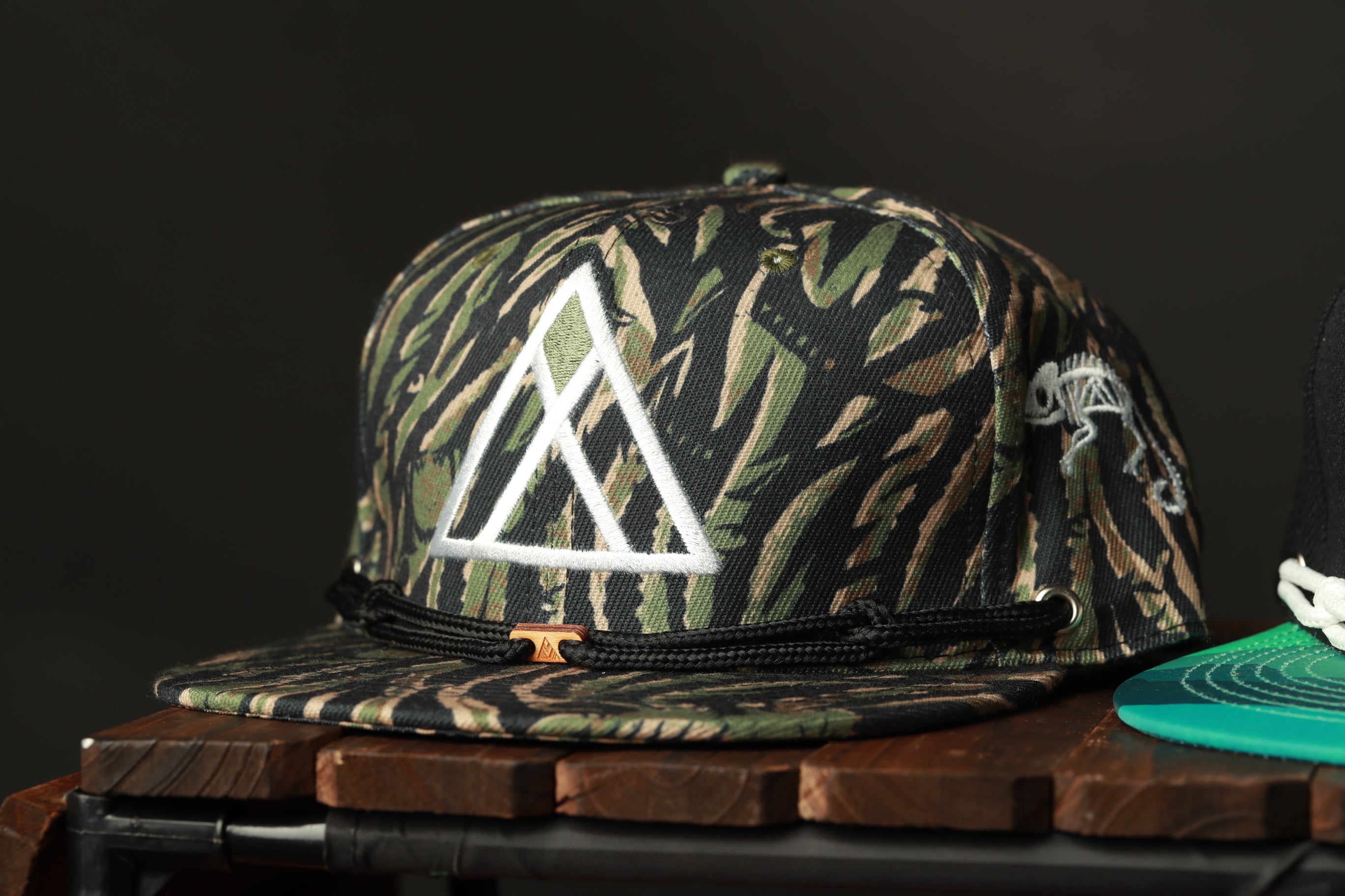A camouflage-patterned baseball cap with a white triangle logo is displayed on a wooden surface.