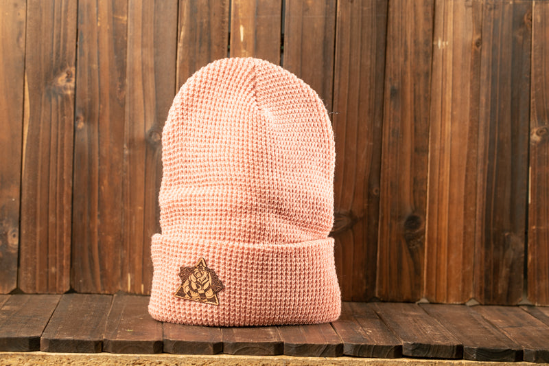 A pink knit beanie with a small logo on the front is displayed against a wooden background.