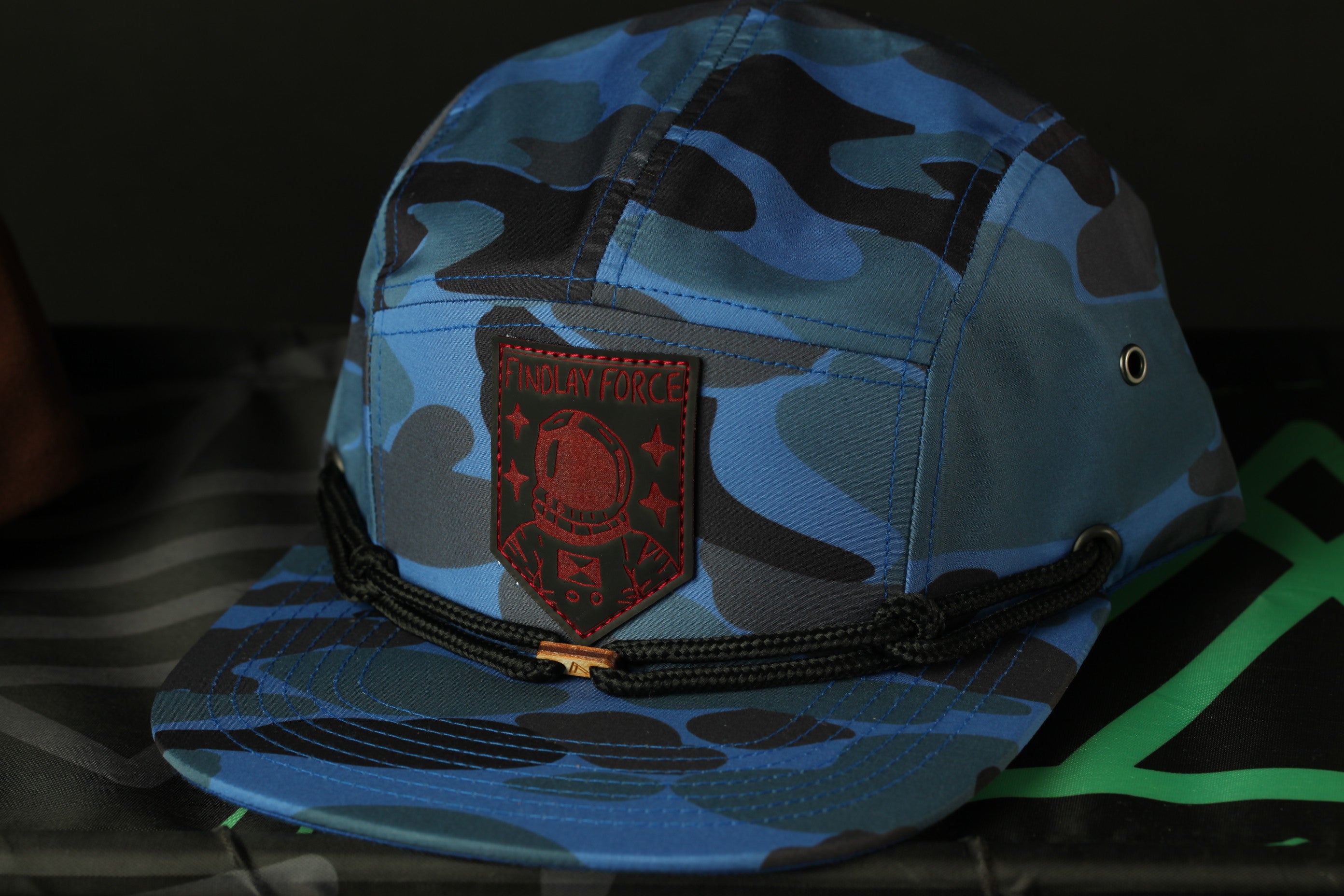 A camouflage baseball cap with a red patch featuring a red helmet and the text "FORD LAYER" printed on it.