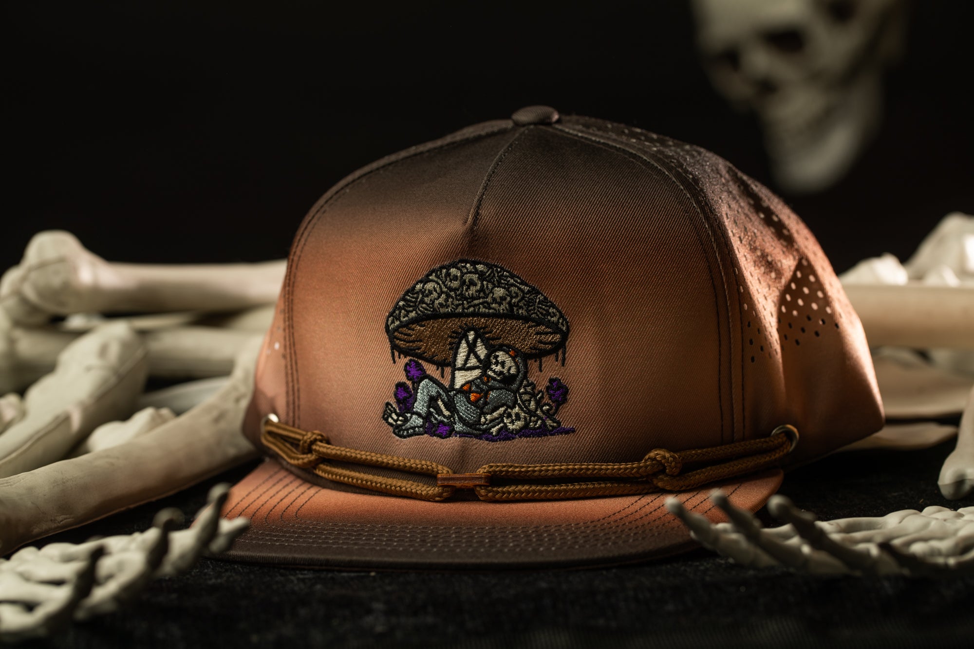A brown baseball cap with an embroidered mushroom design sits on a surface surrounded by skeletal bones.