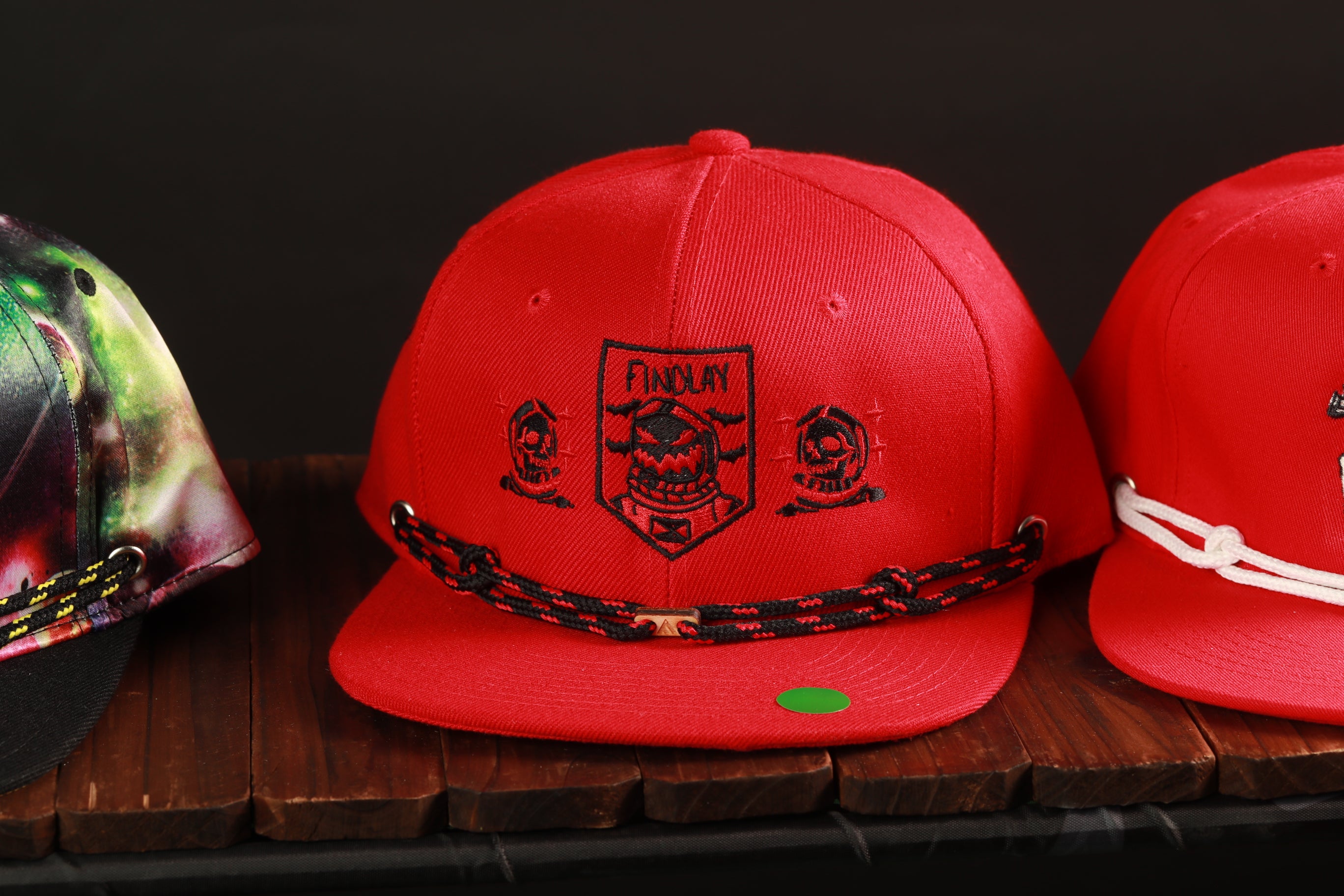 A red baseball cap with a skull and crossbones design, featuring the text "FINALLY" on the front, is displayed on a wooden surface.