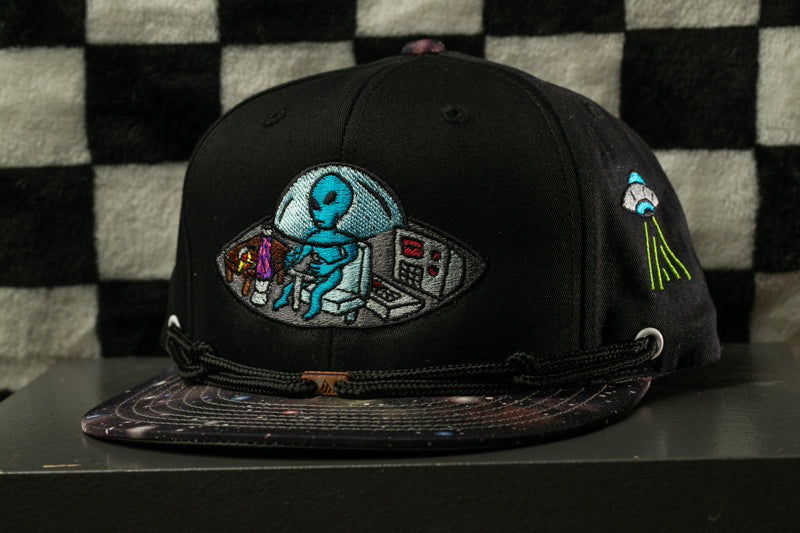 A black baseball cap with a colorful alien embroidered on it, sitting in a space-themed cockpit.