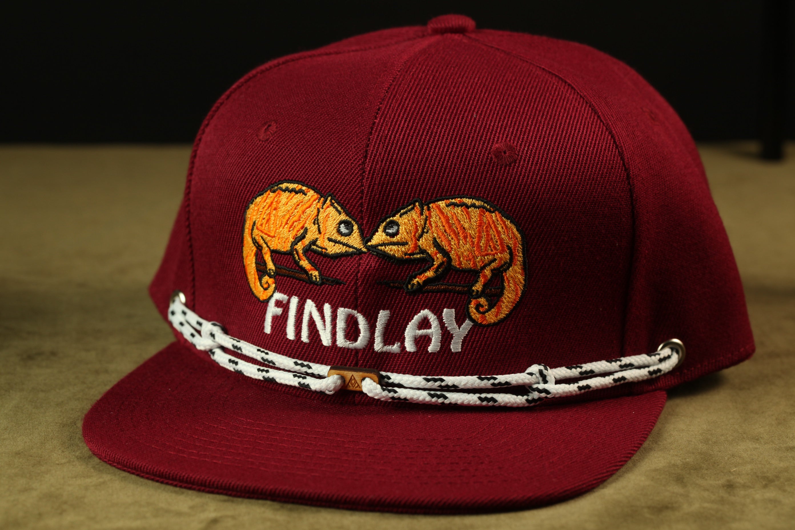 A maroon baseball cap with a white string and a logo of two orange chameleons.