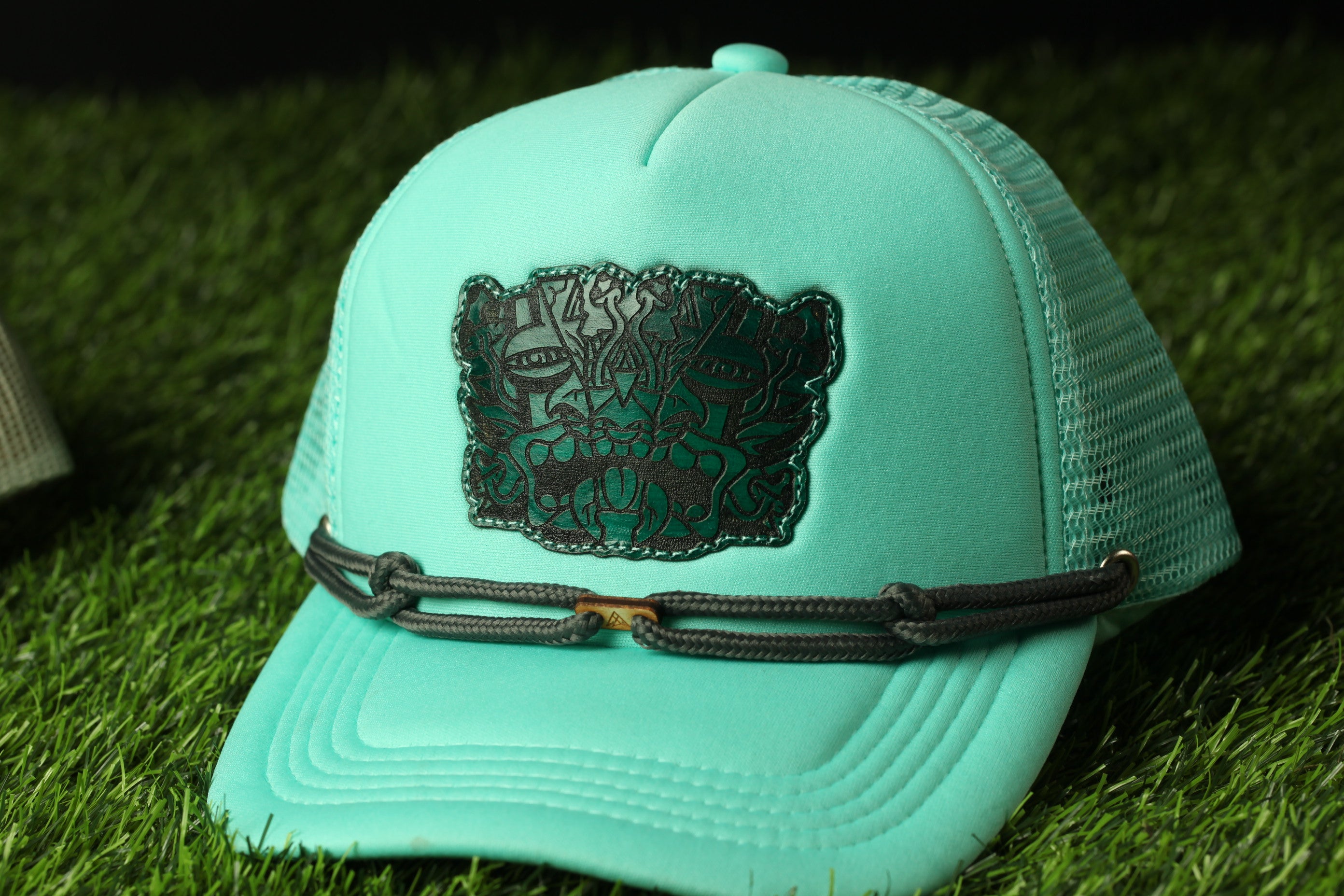 A turquoise baseball cap with a black and green embroidered logo on the front, resting on a grassy surface.