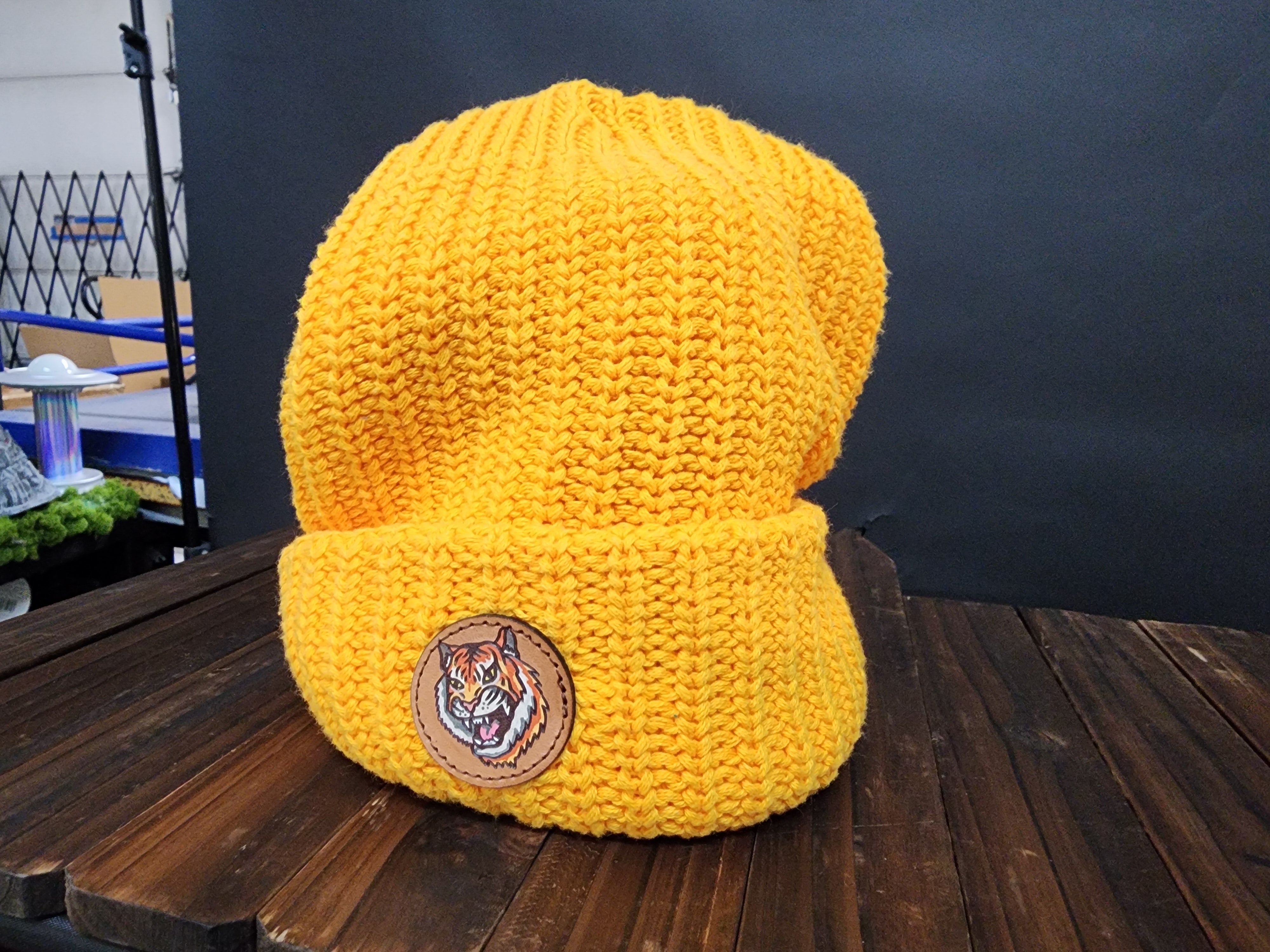 A bright yellow knit beanie with a tiger logo is displayed on a wooden surface.