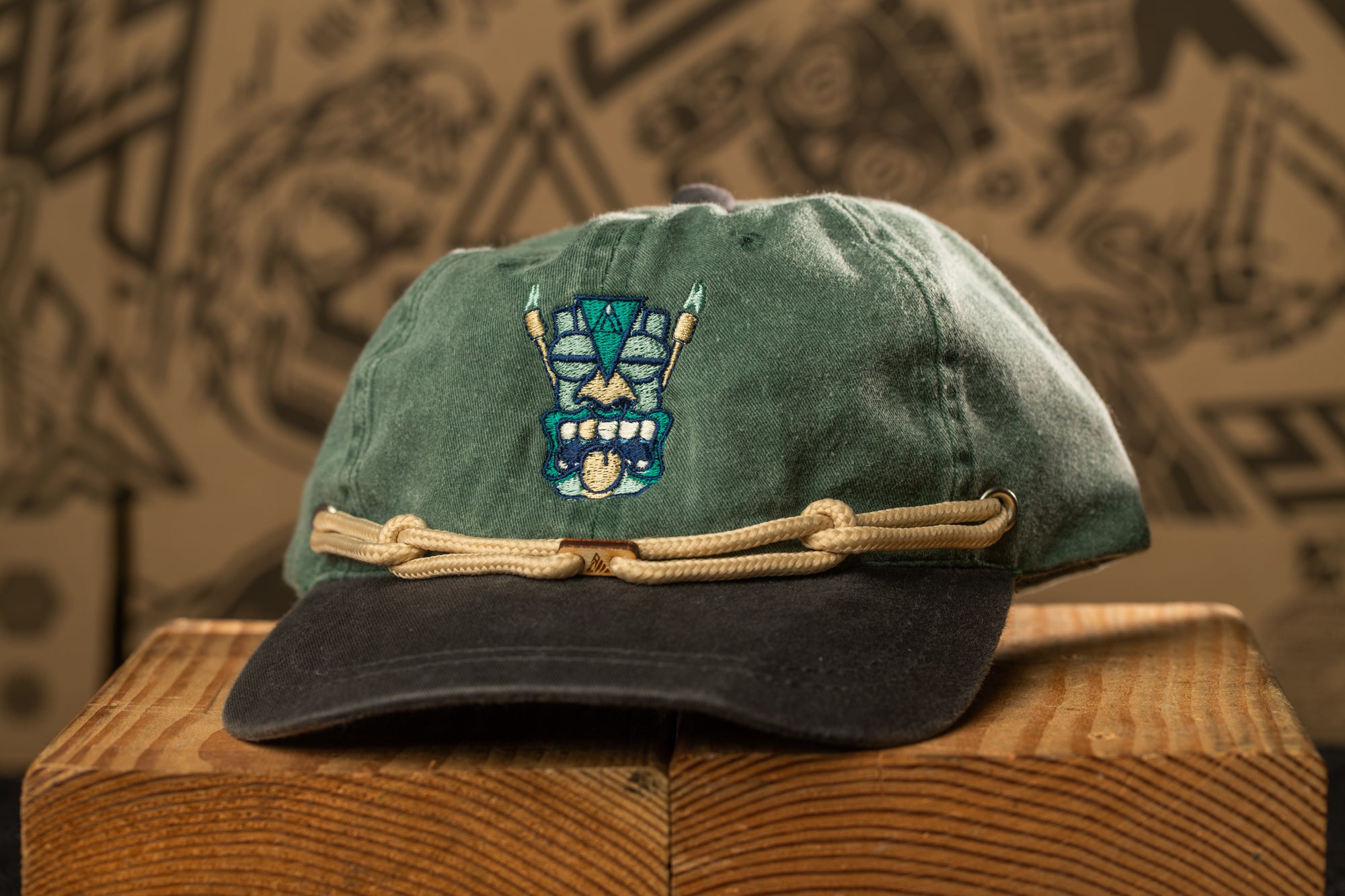 A green baseball cap with a tiki mask design on the front, placed on a wooden surface against a blurred background.