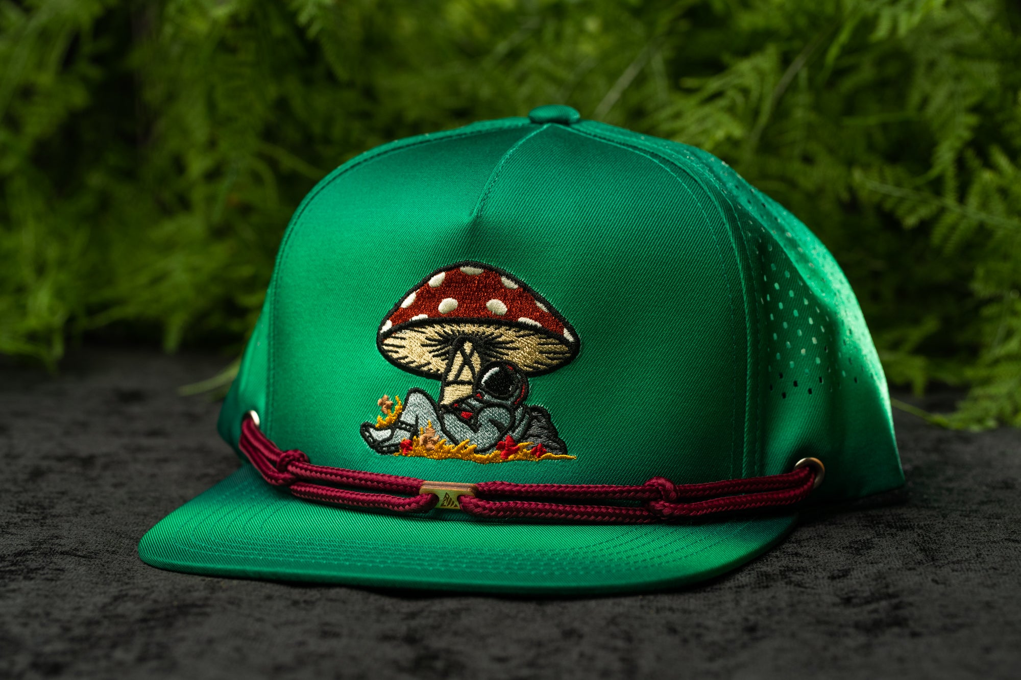 A green baseball cap with a mushroom design and a red string around the brim, placed on a dark surface with green foliage in the background.