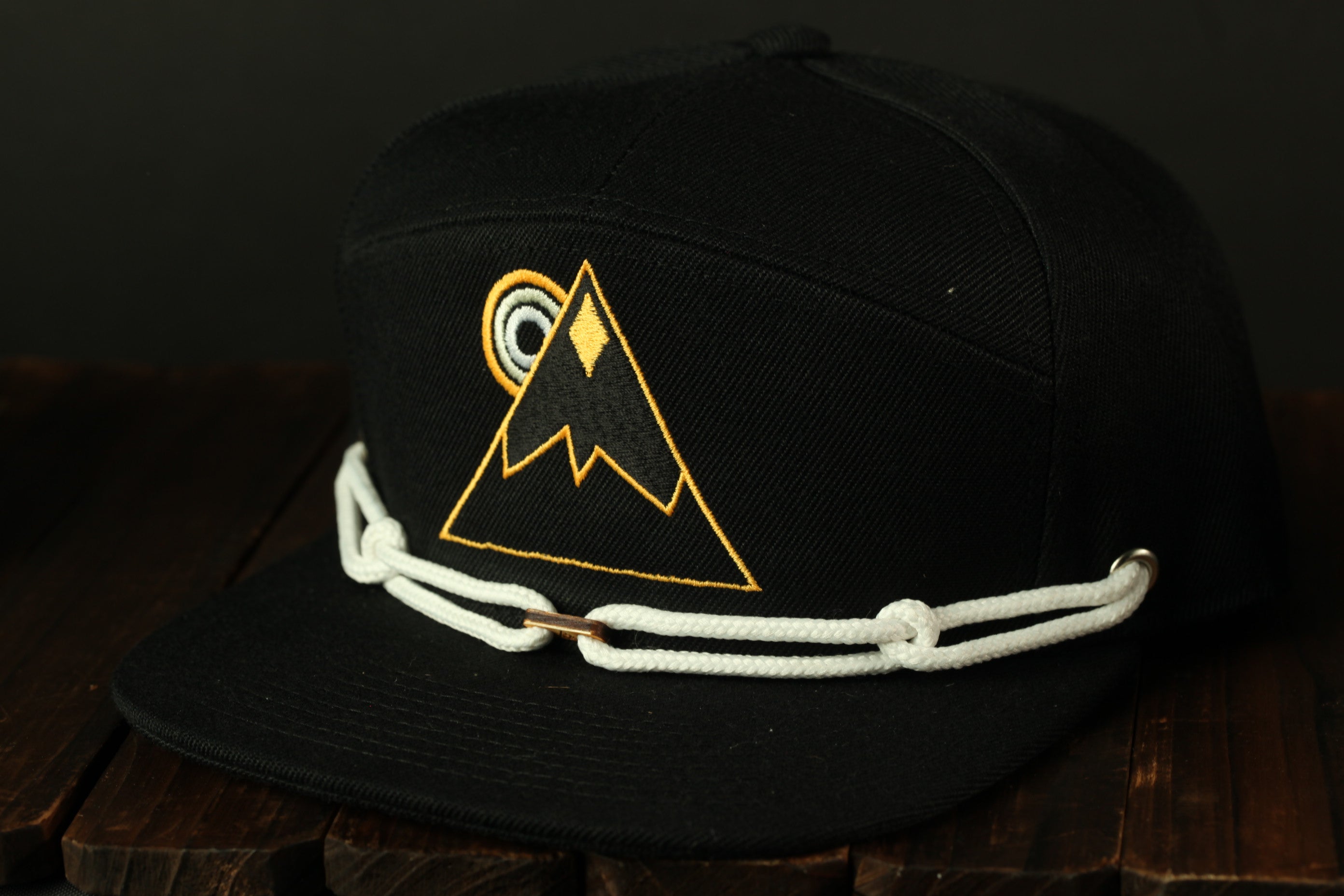A black baseball cap with a yellow mountain logo and white rope strap.