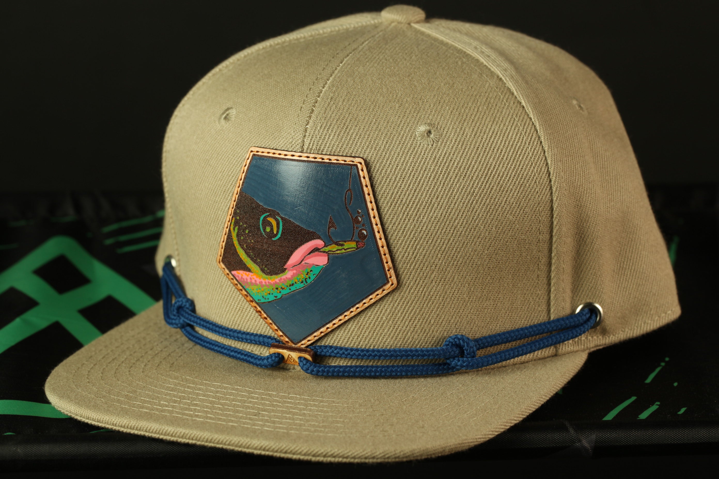 A beige baseball cap with a blue and orange patch featuring a fish design, and a blue string or strap.
