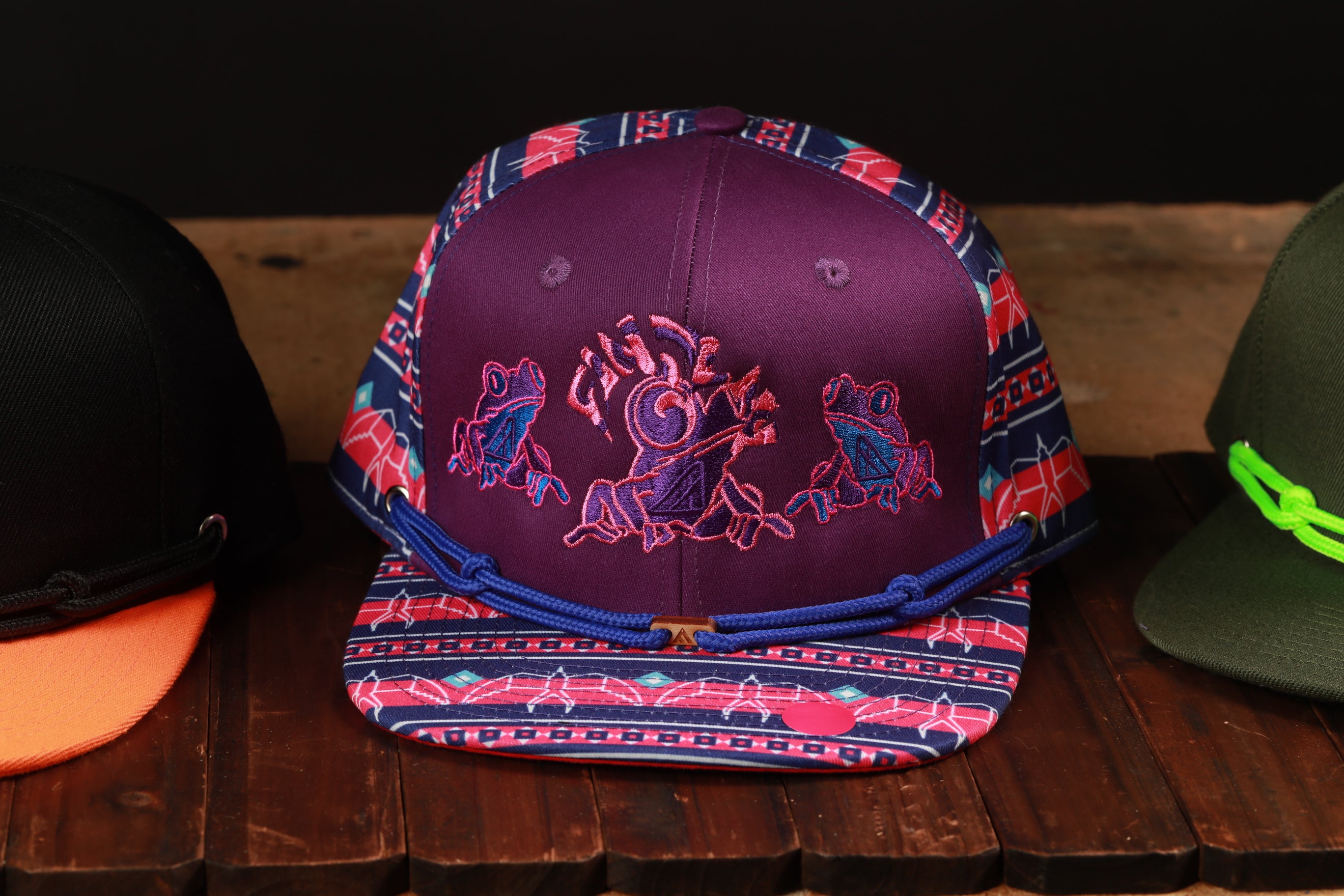 A purple baseball cap with a colorful, embroidered design featuring frogs and other wildlife, displayed on a wooden surface.