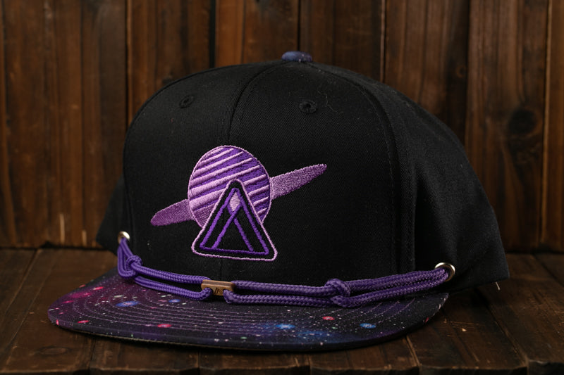 A black baseball cap with a purple and purple galaxy-themed design on the front, sitting on a wooden surface.