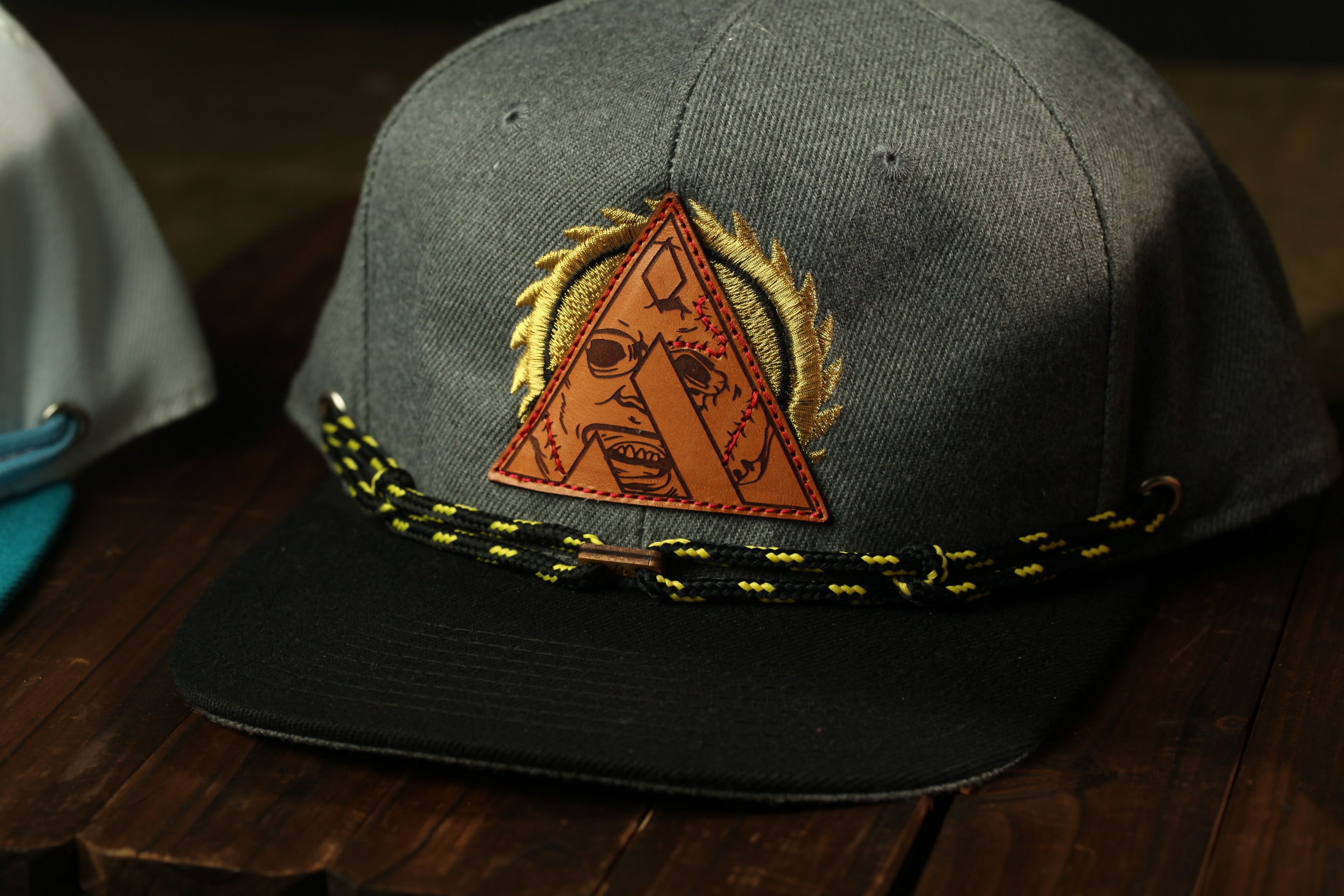 A gray baseball cap with a triangular patch featuring a skull and crossbones design, and a black and yellow braided strap.