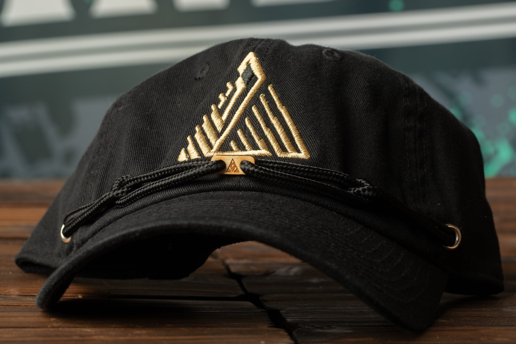 A black baseball cap with a gold geometric pattern on the front, resting on a wooden surface.