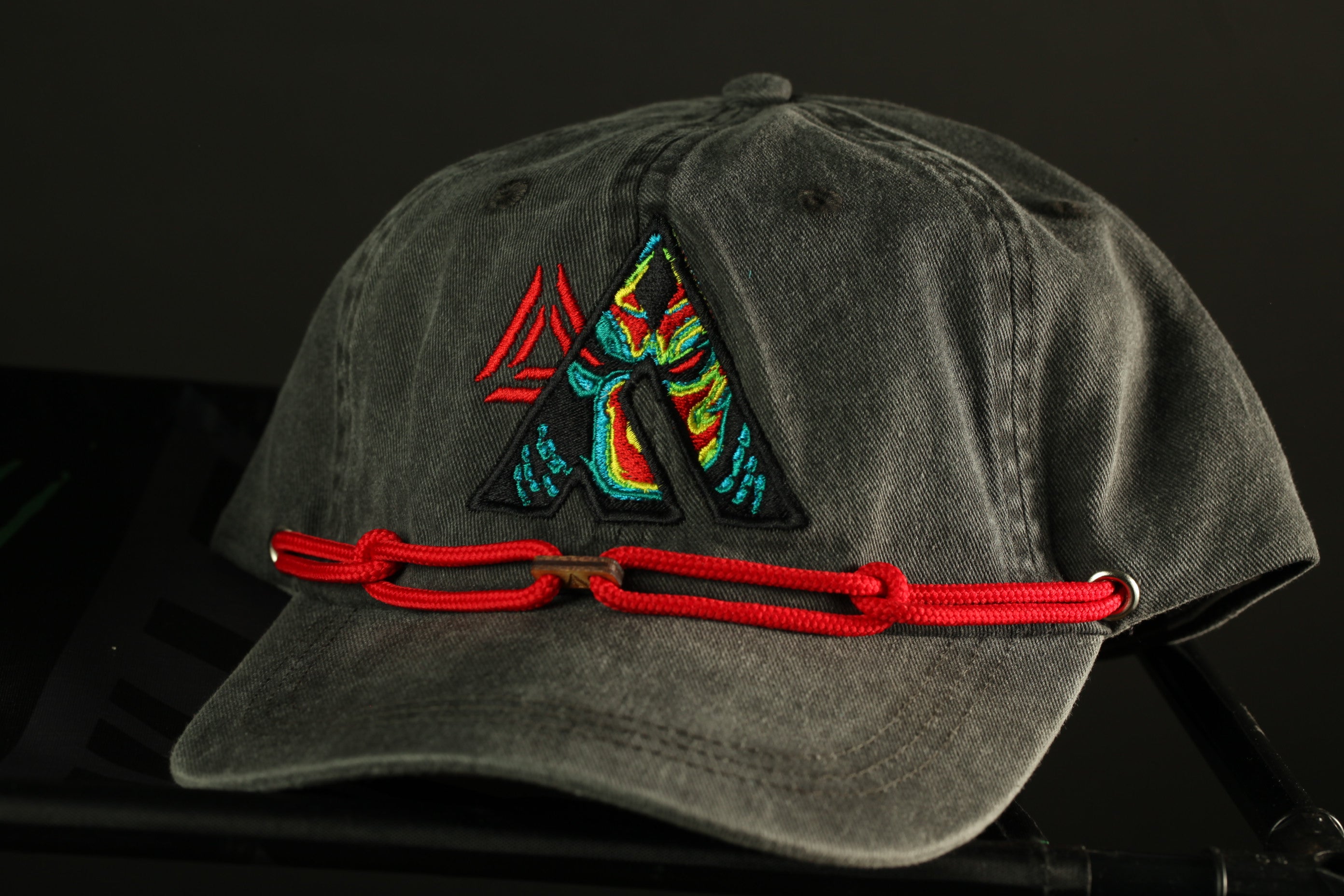 A gray baseball cap with a colorful, embroidered logo on the front, and a red chain strap.