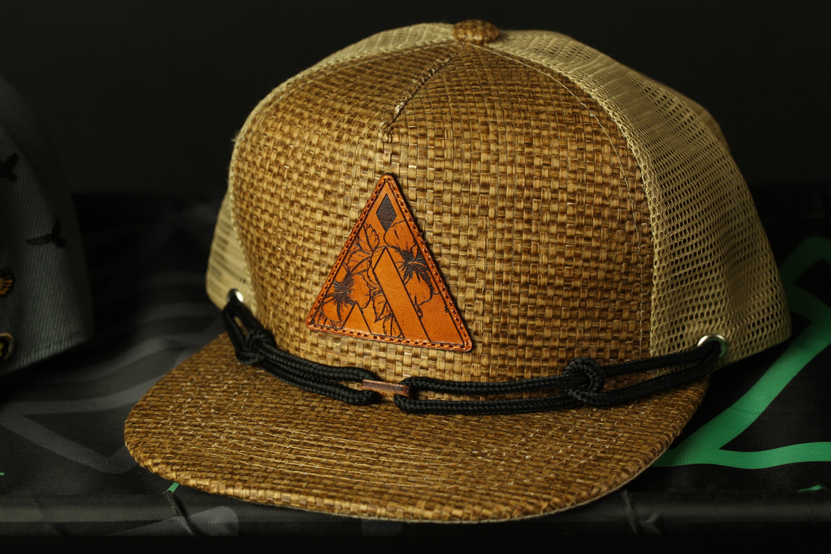 A brown woven baseball cap with a black lace detail and a triangle-shaped patch on the front.