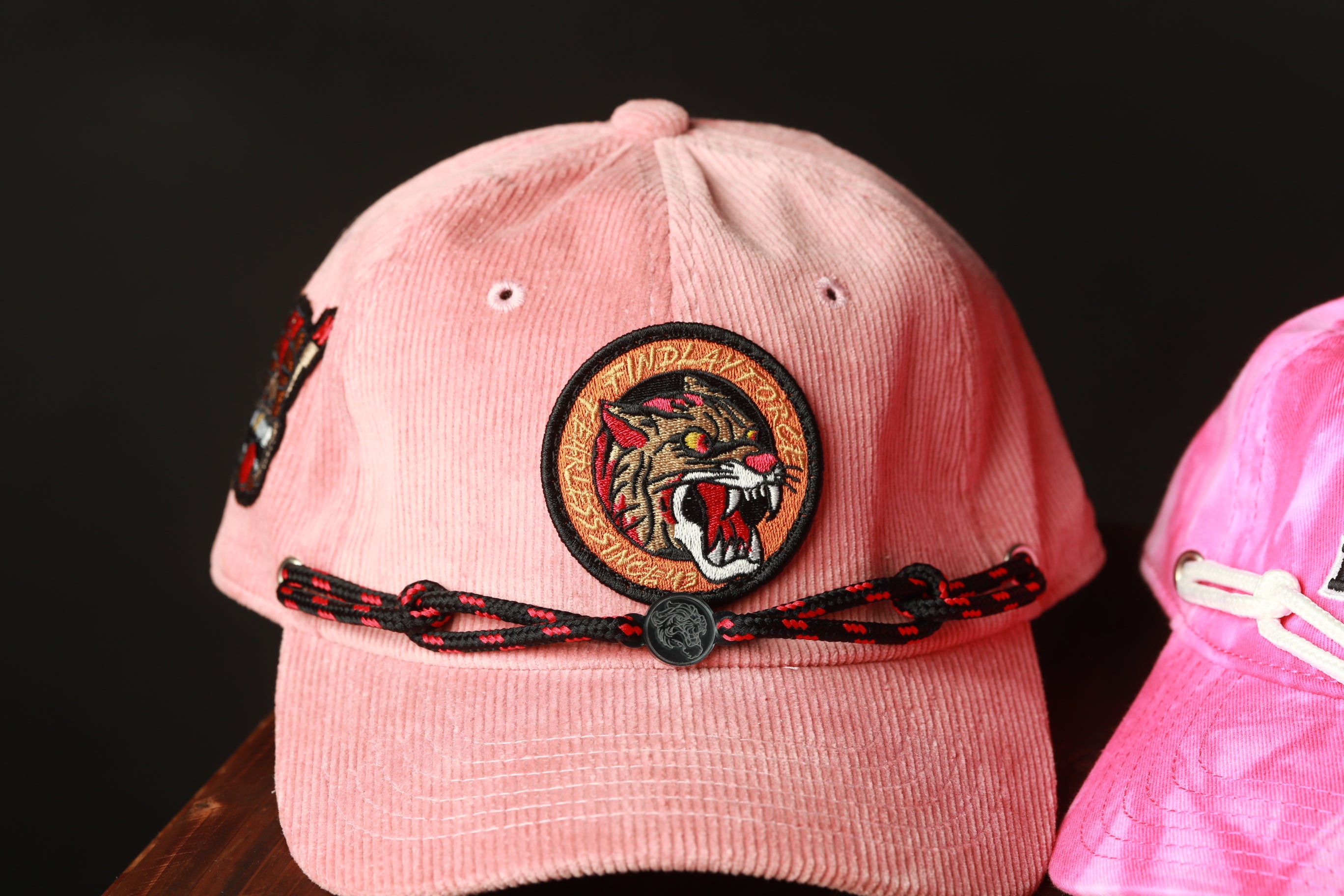 A pink baseball cap with a black and red braided strap and a circular patch featuring a tiger's face.