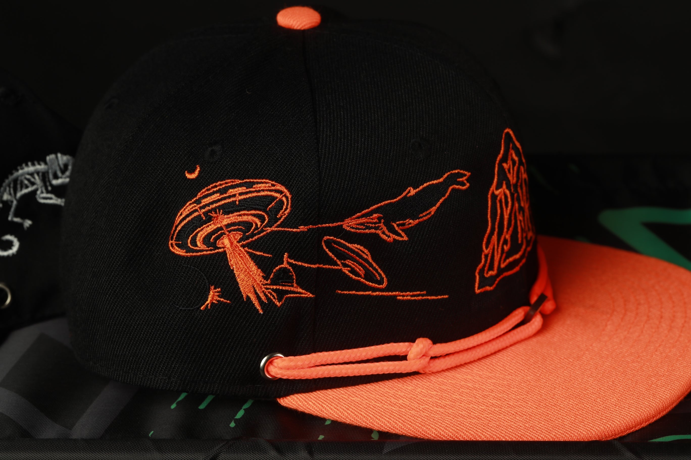 A black baseball cap with an orange design on it, featuring a stylized illustration of a fish and a mushroom.