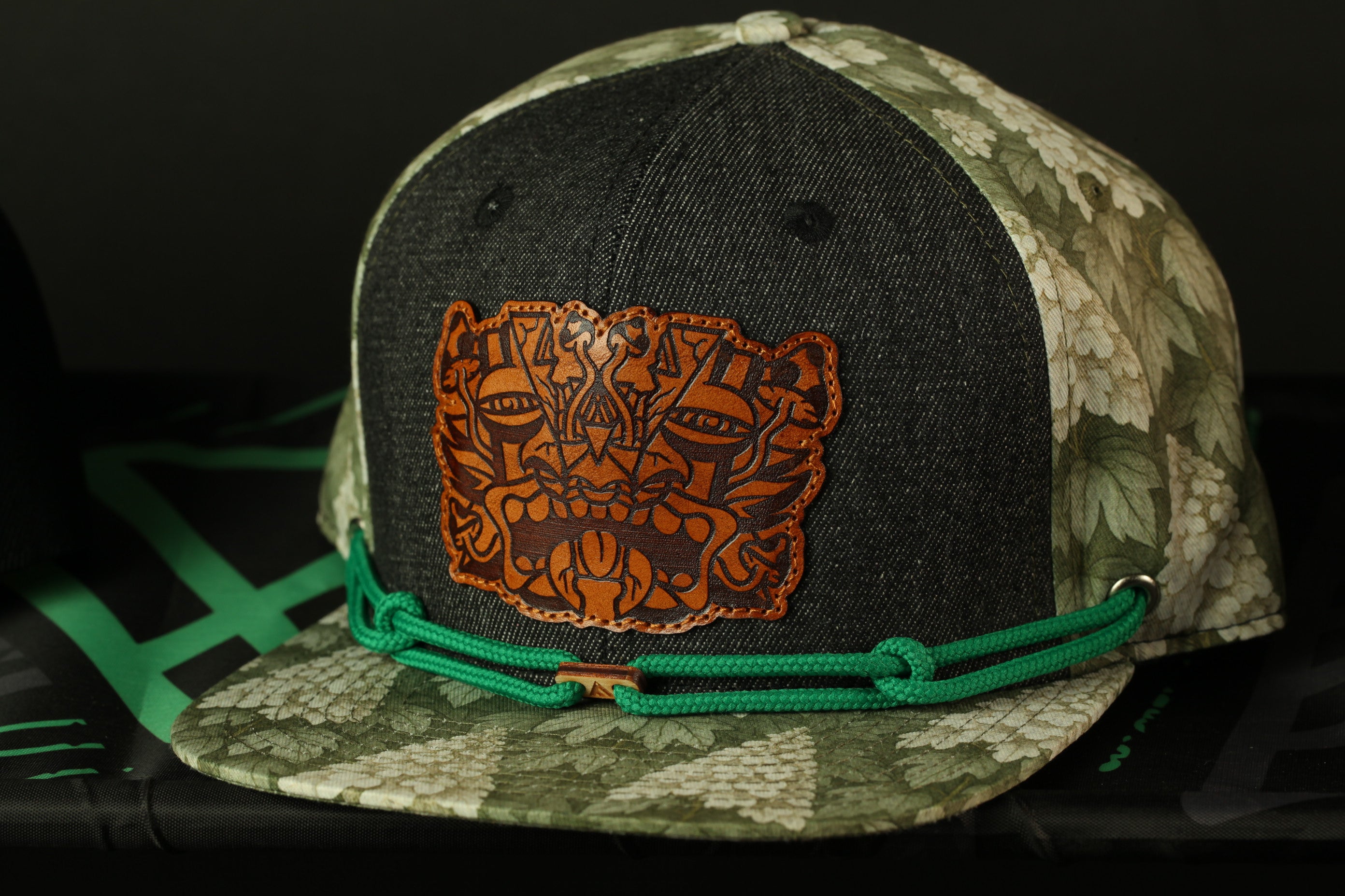 A black and green camouflage baseball cap with a brown leather patch featuring an abstract design on the front.