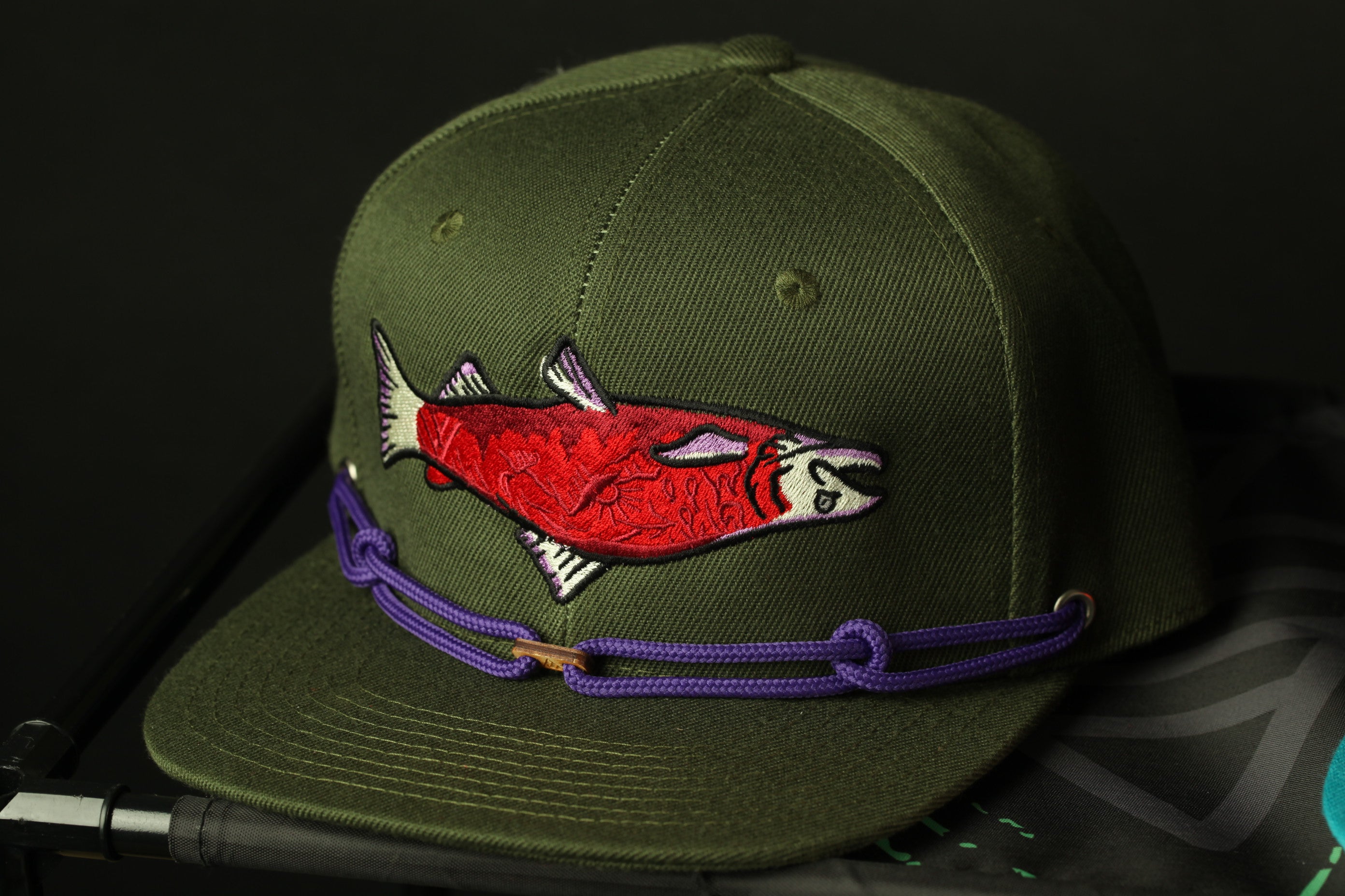 A green baseball cap with a purple string and a red fish embroidered on it.