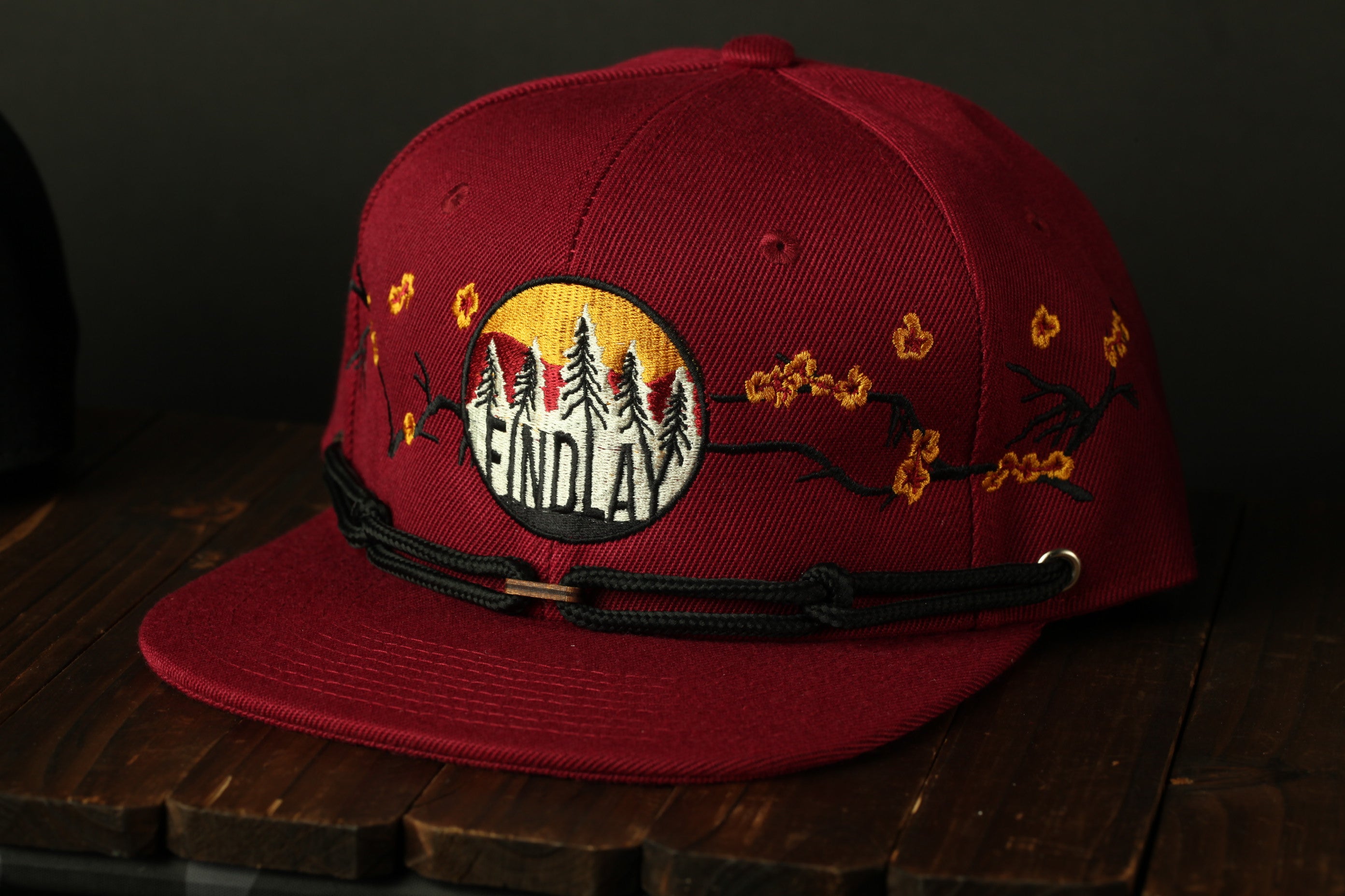 A red baseball cap with a black band and embroidered designs, including trees and flowers, on the front.