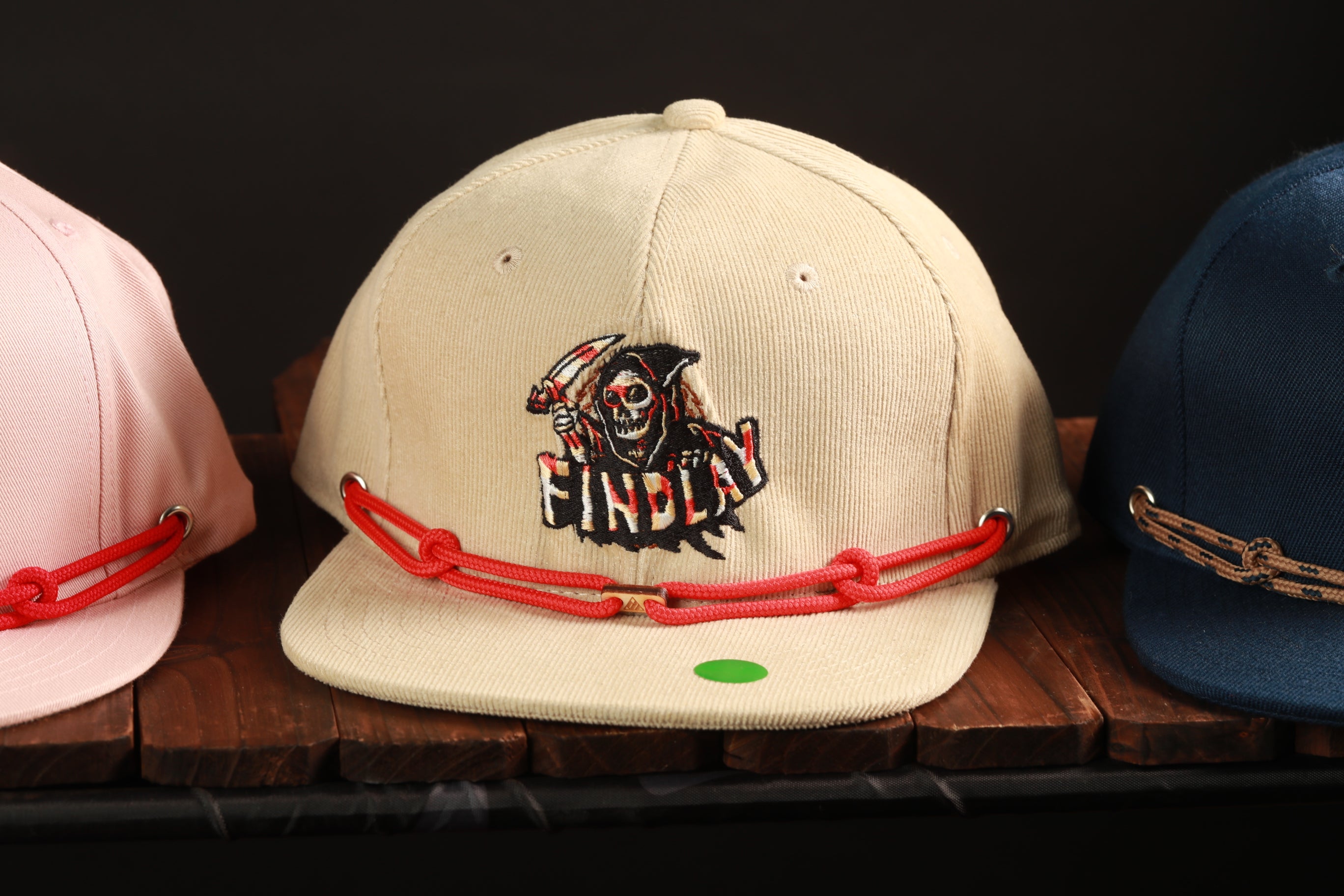 A beige baseball cap with a skull and crossbones design and the word "FINDlay" printed on it, along with a red string and a green tag.
