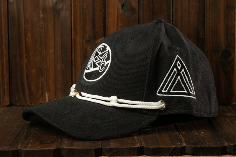 A black baseball cap with white embroidered logos and a white rope strap is displayed on a wooden surface.