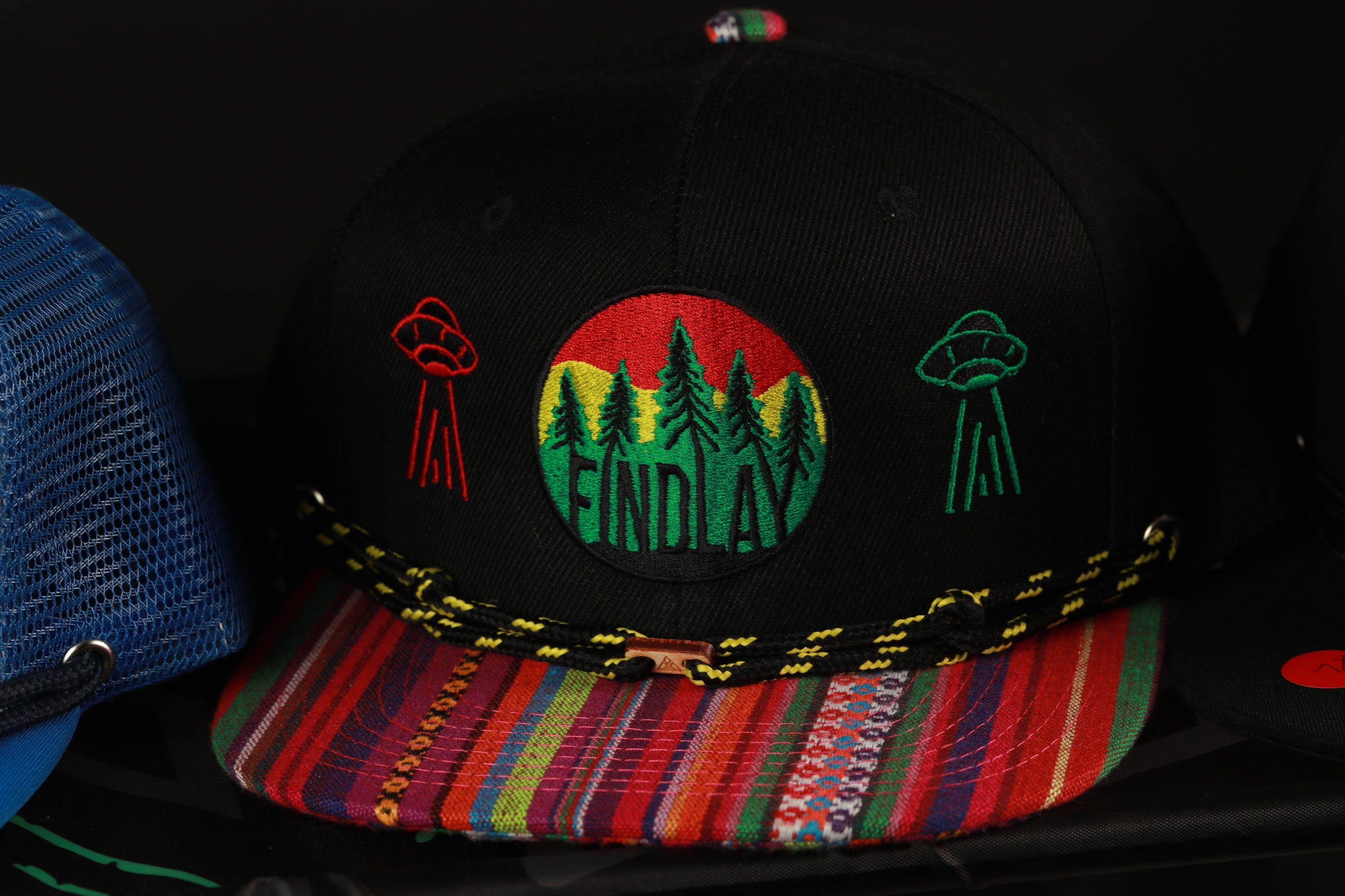 A black baseball cap with colorful stripes and embroidered designs, including a circular patch with the word "FIND" and two alien-like figures.