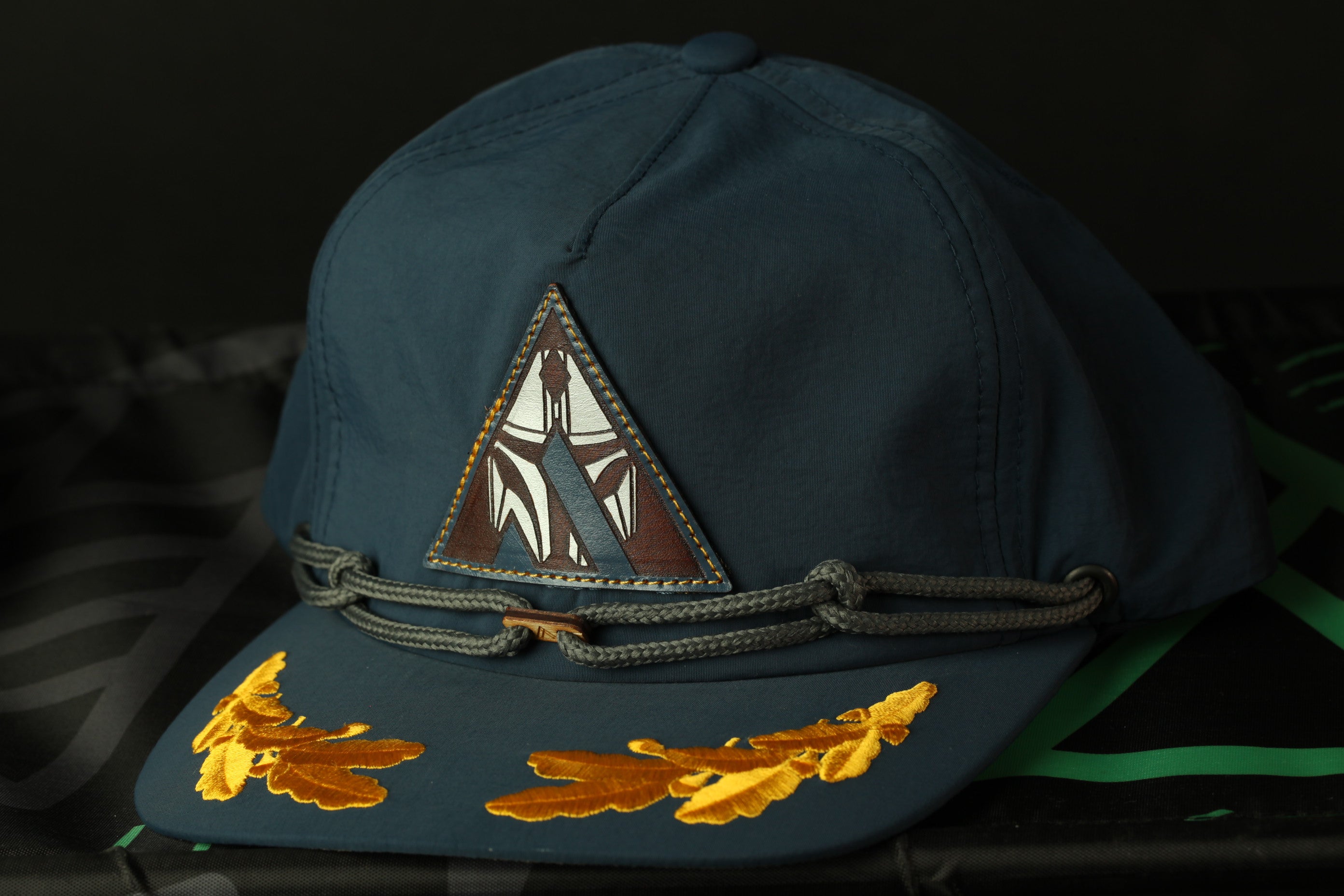 A navy blue baseball cap with a triangular patch and gold leaf embroidery on the brim.
