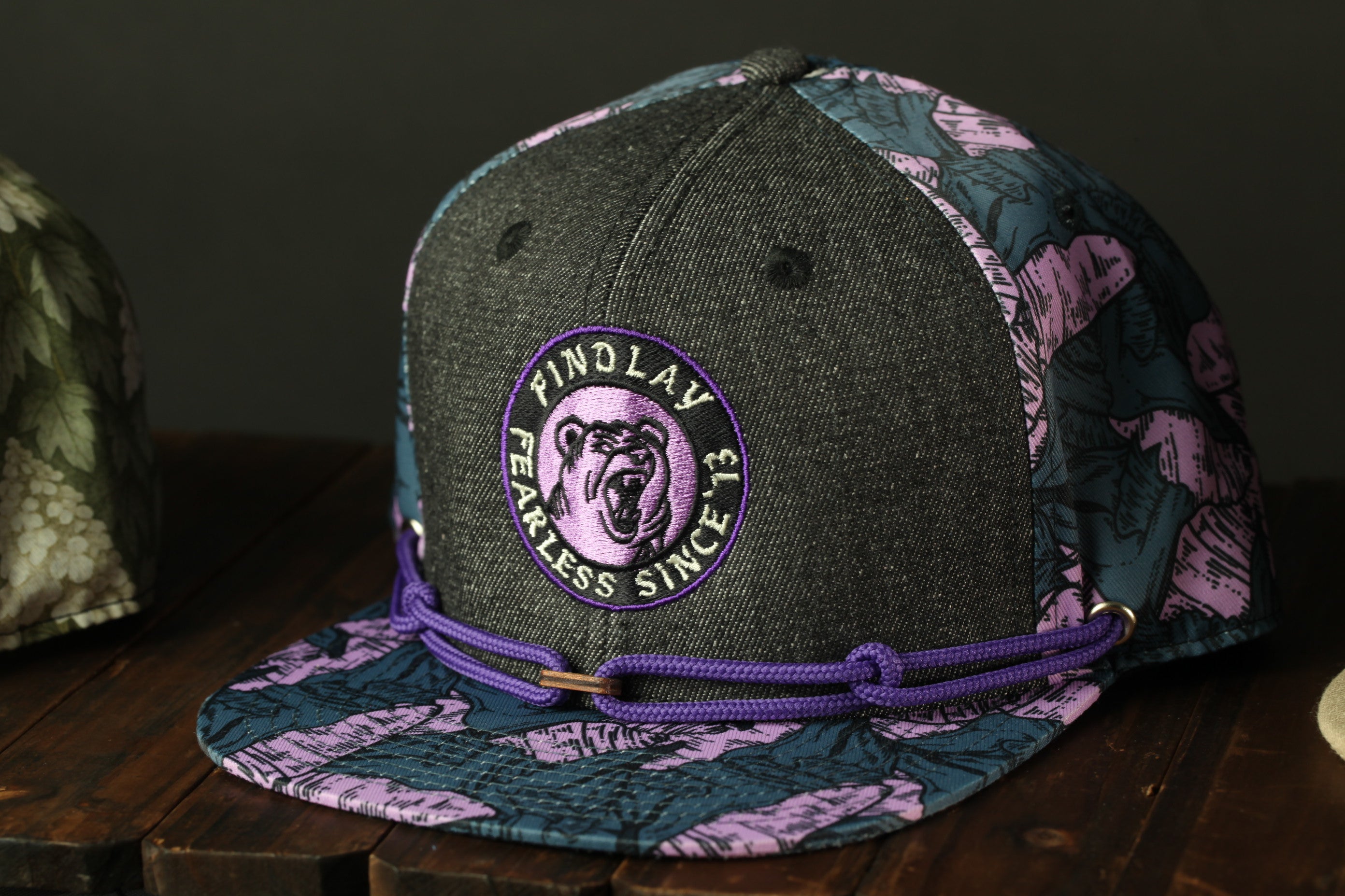 A black and purple baseball cap with a purple and pink floral pattern and a purple ribbon around the brim.