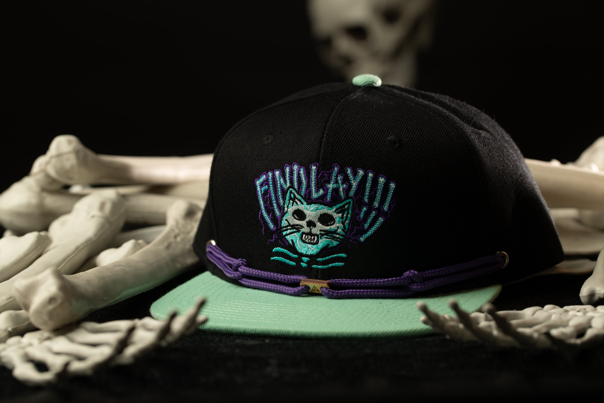 A black baseball cap with a cat design and the text "FIND LAYY!!!" is displayed on a table, surrounded by white bones.