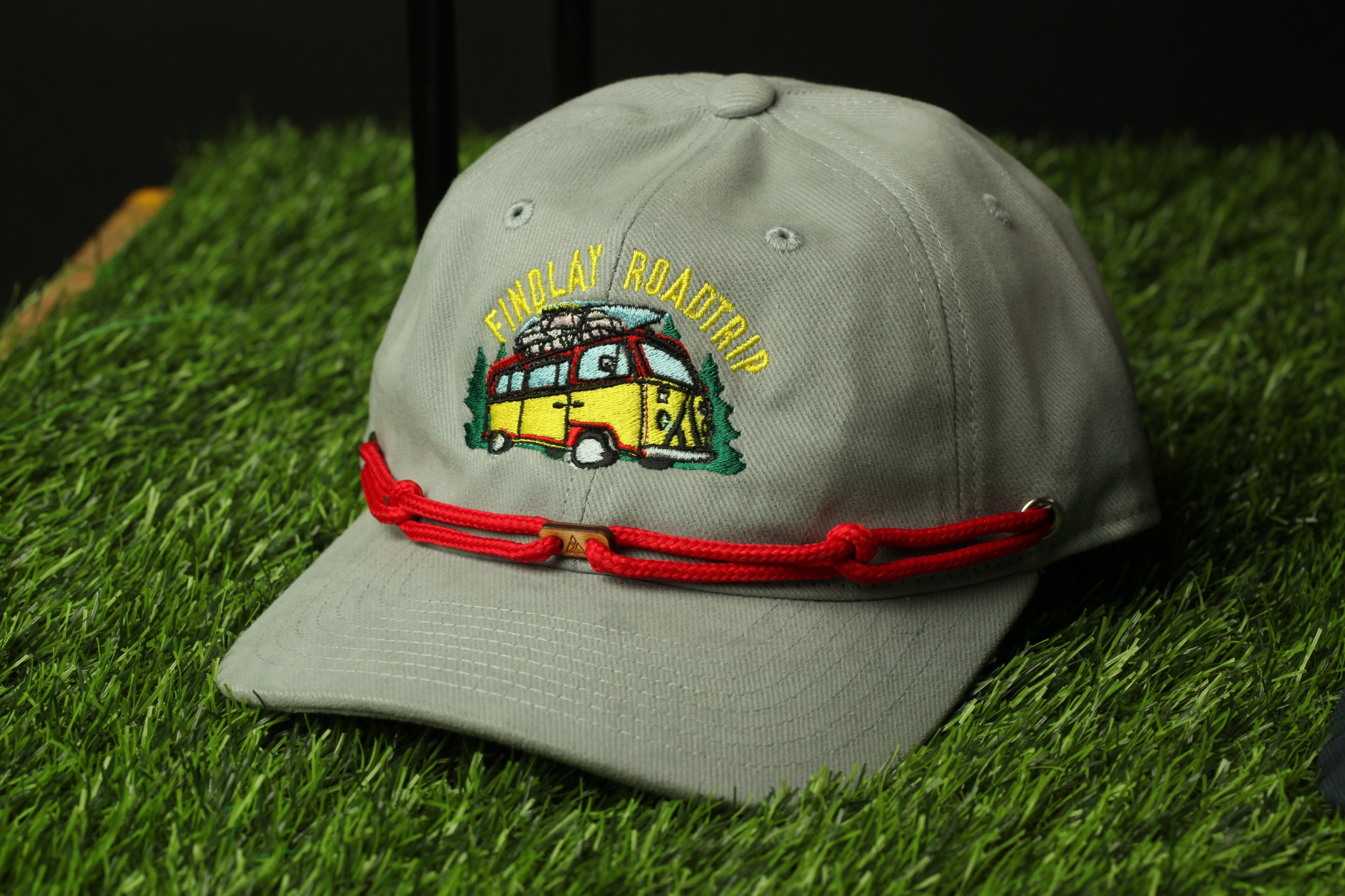 A gray baseball cap with a red string and a yellow and green embroidered design on the front, sitting on a green surface.