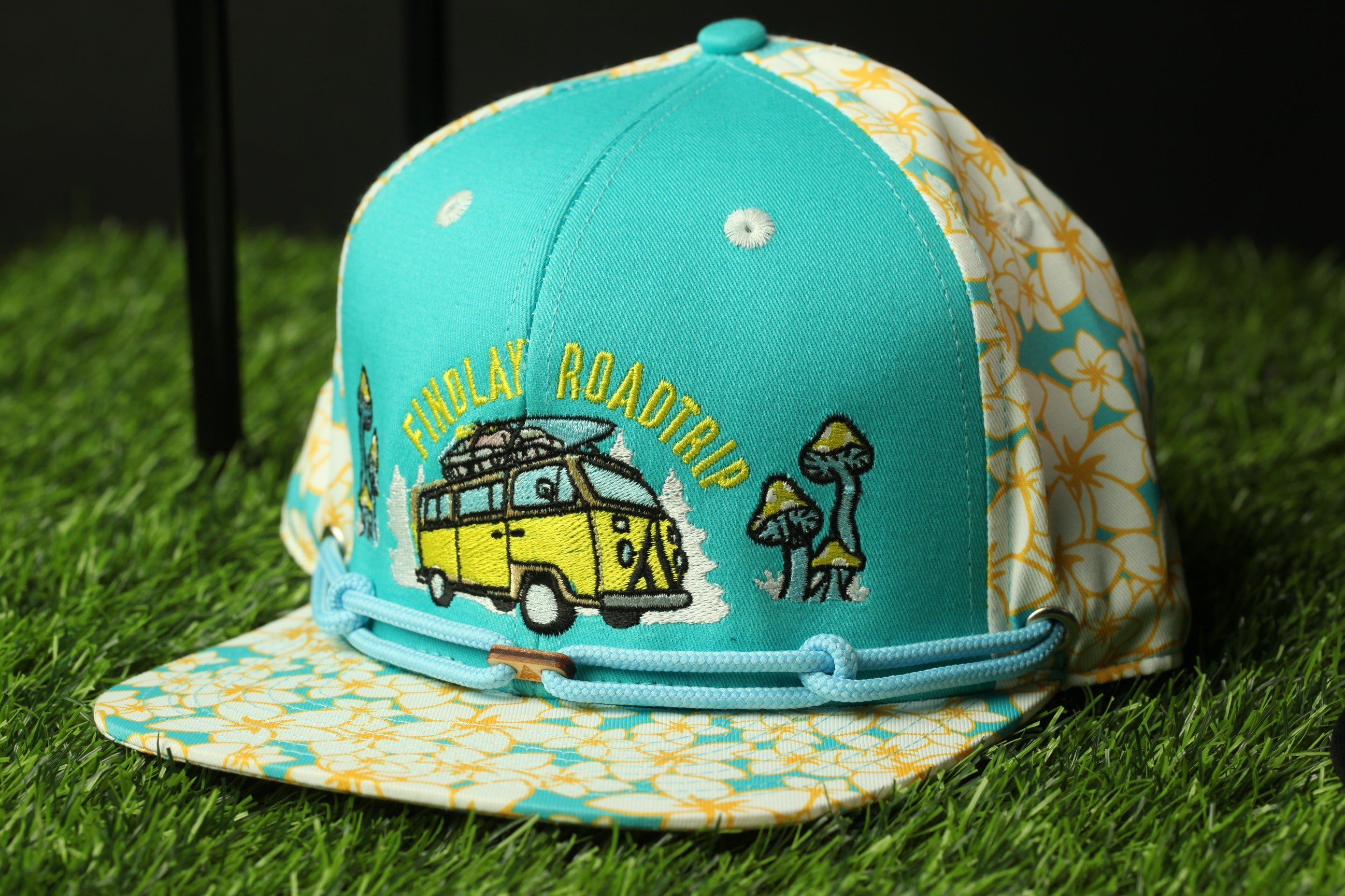 A turquoise baseball cap with a floral pattern and a yellow van design, sitting on a grassy surface.