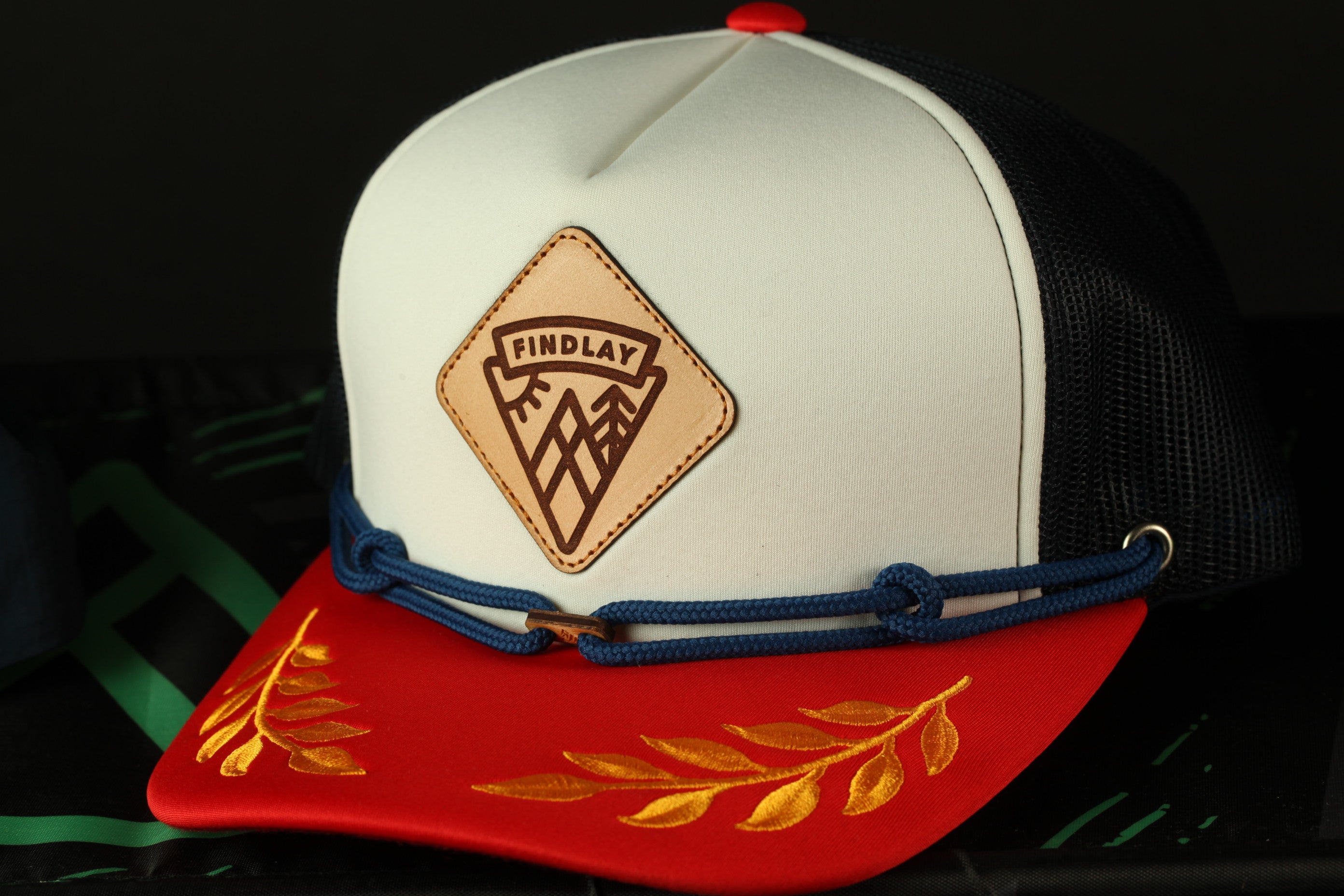 A white baseball cap with a red brim and a leather patch on the front, featuring a gold leaf design.