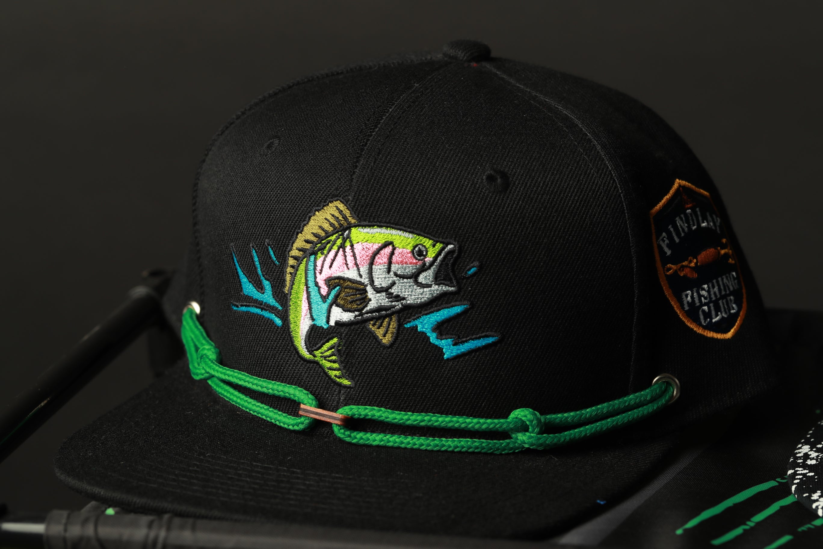 A black baseball cap with a colorful fish embroidered on it, featuring a green chain around the brim.