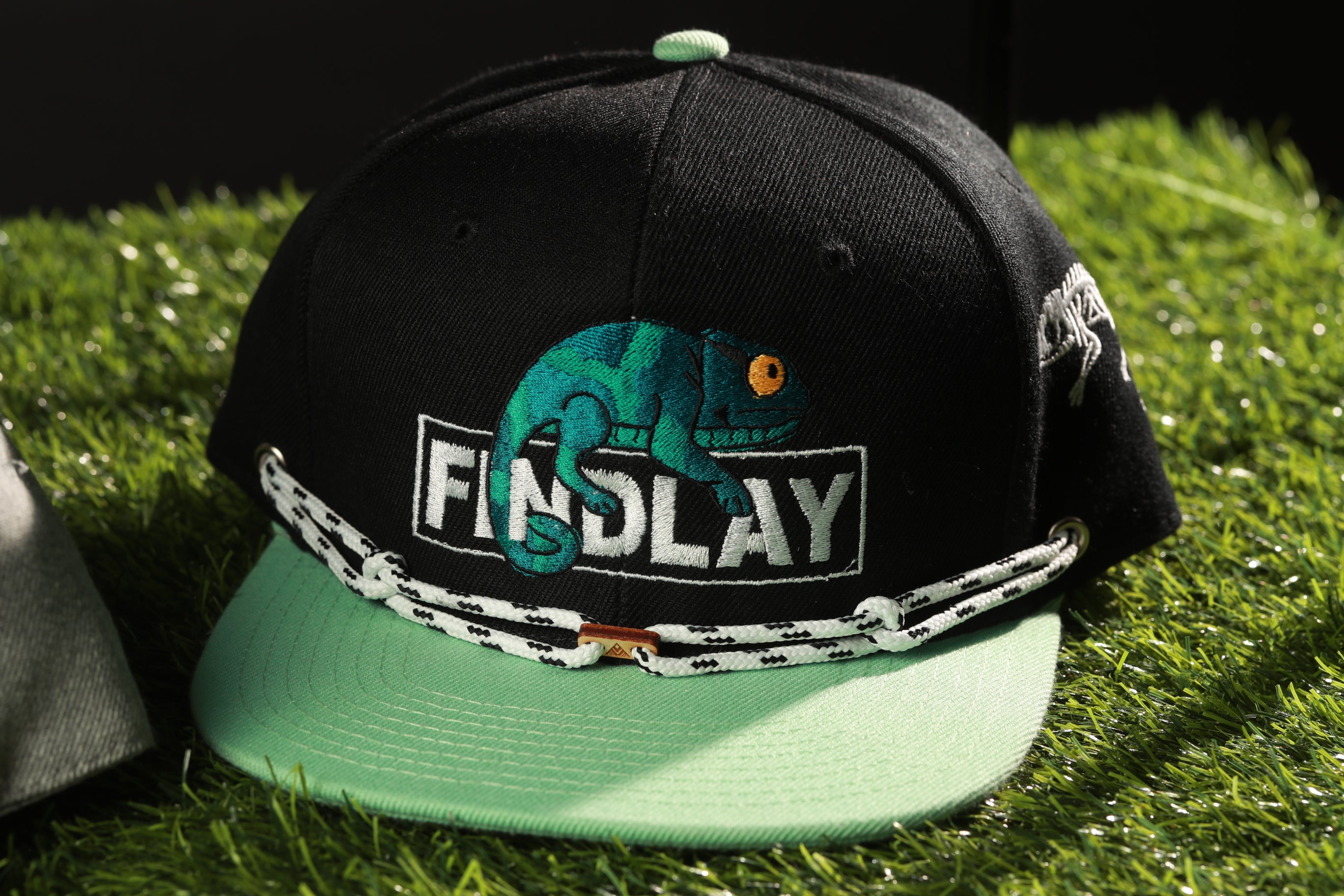 A black baseball cap with a green brim and a white and green chain strap, featuring a chameleon design and the word "FINDlay" printed on it.