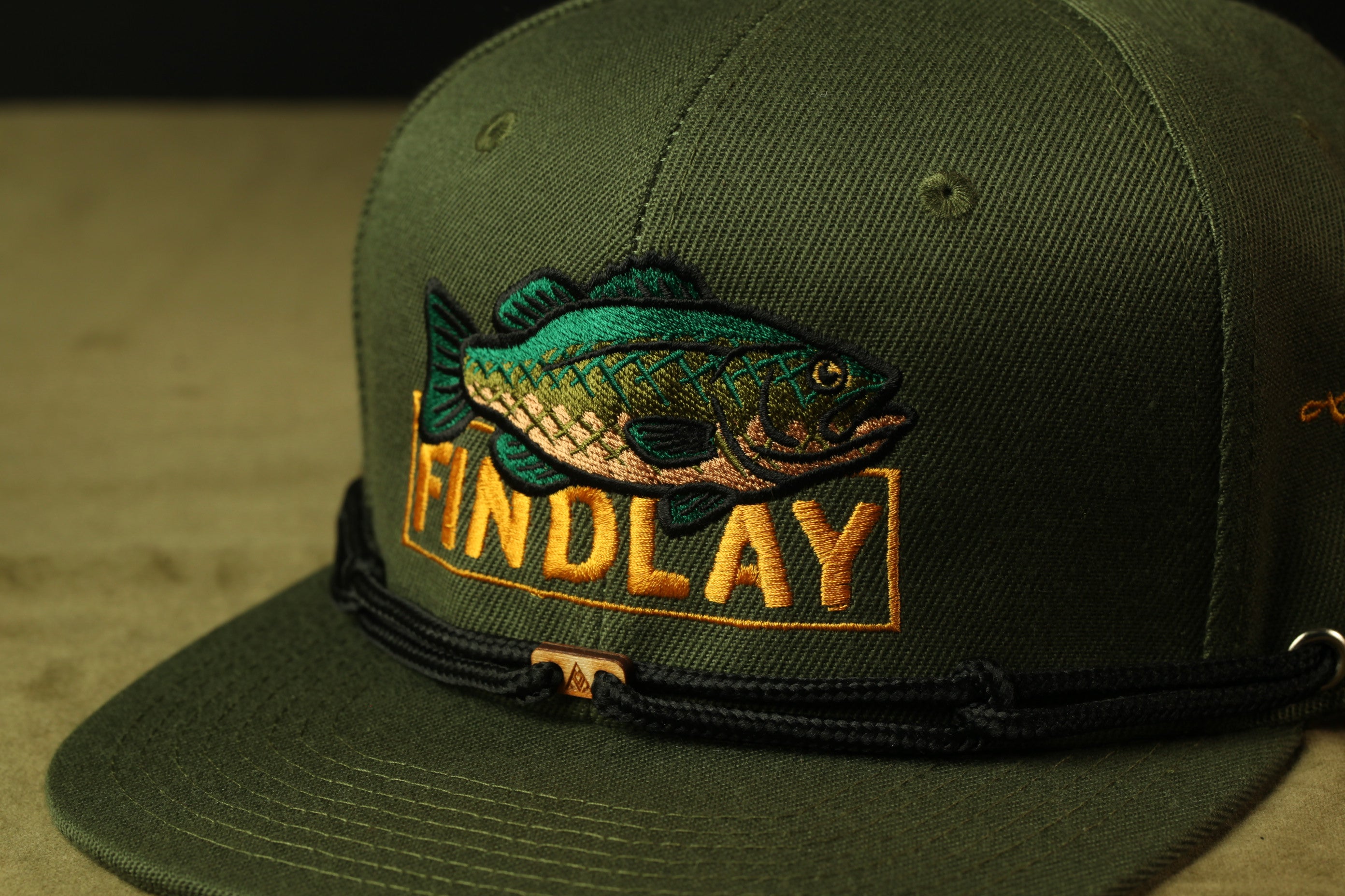 A close-up of a green baseball cap with a fish embroidered on it, featuring the word "FINDlay" in orange text.