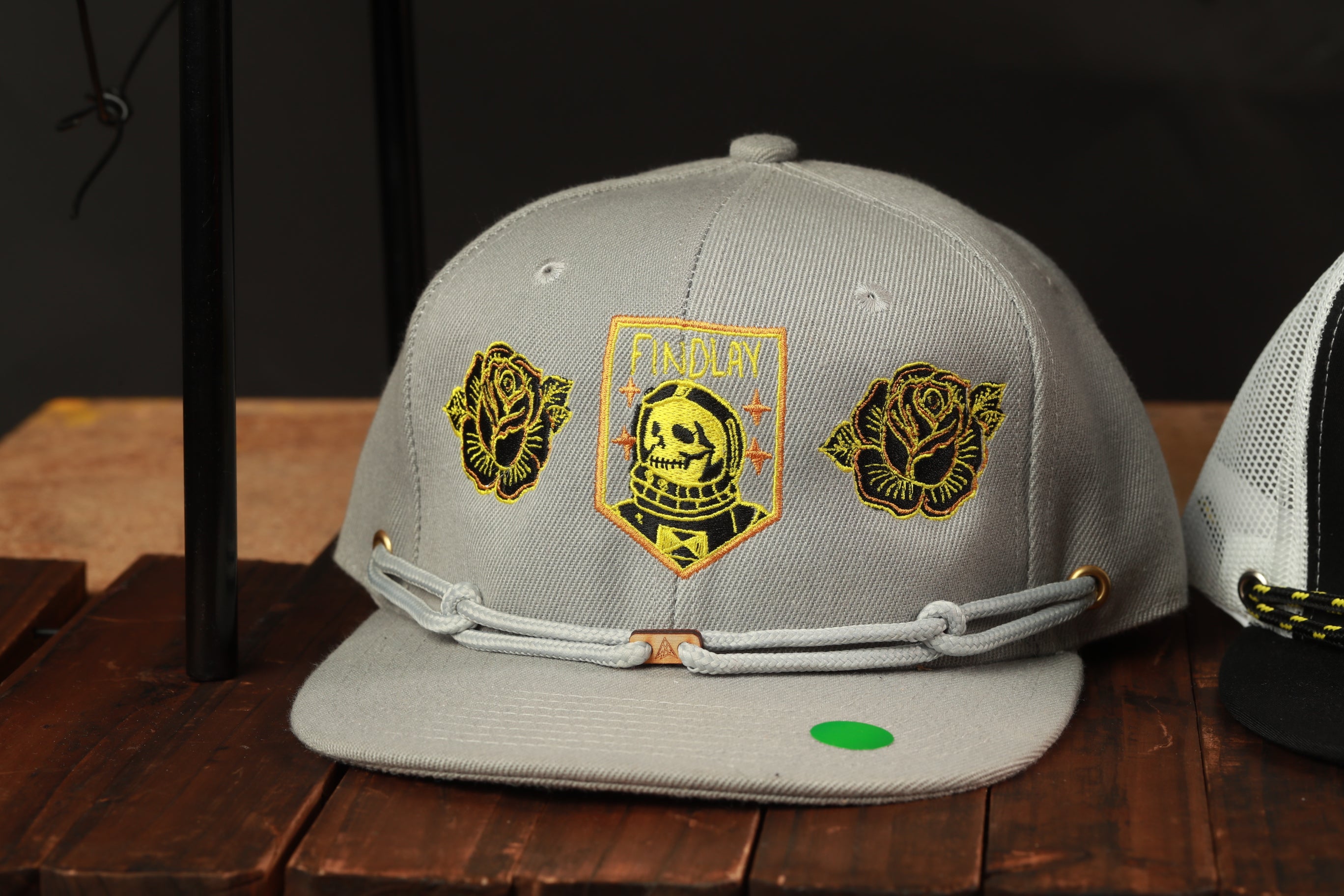 A gray baseball cap with various embroidered designs and patches, including a skull and rose motif, is displayed on a wooden surface.