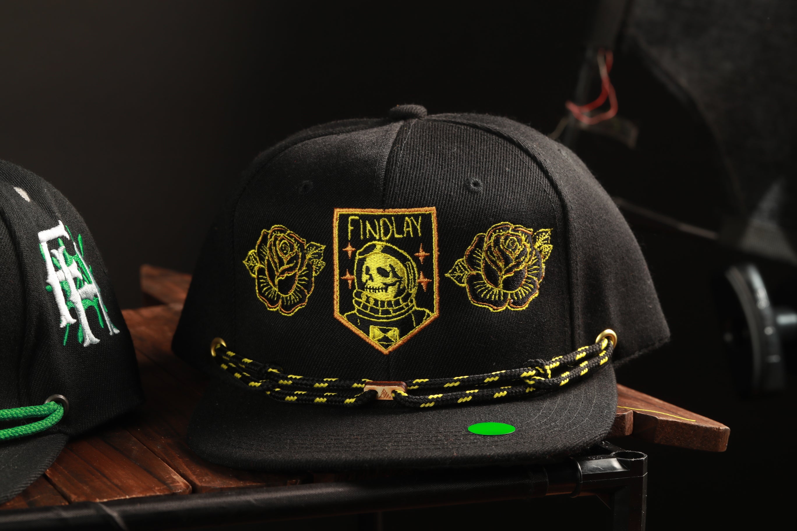 A black baseball cap with gold embroidered designs and text, including a skull and rose motif, is displayed on a wooden surface.